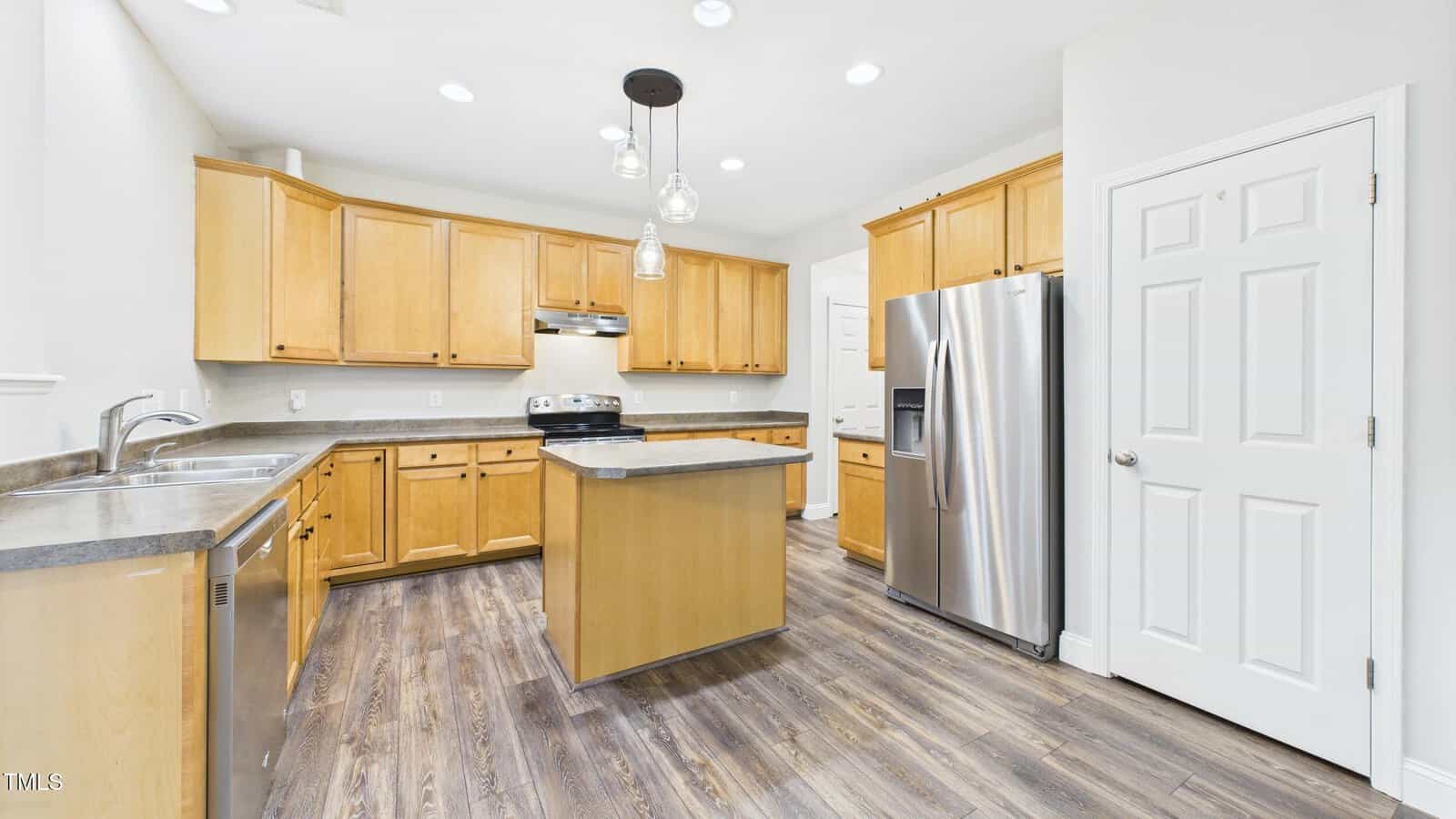 Spacious modern kitchen with light wood cabinets, stainless steel appliances, gray countertops, pendant lights, and wood-look flooring. There is an island in the center and a pantry door on the right.