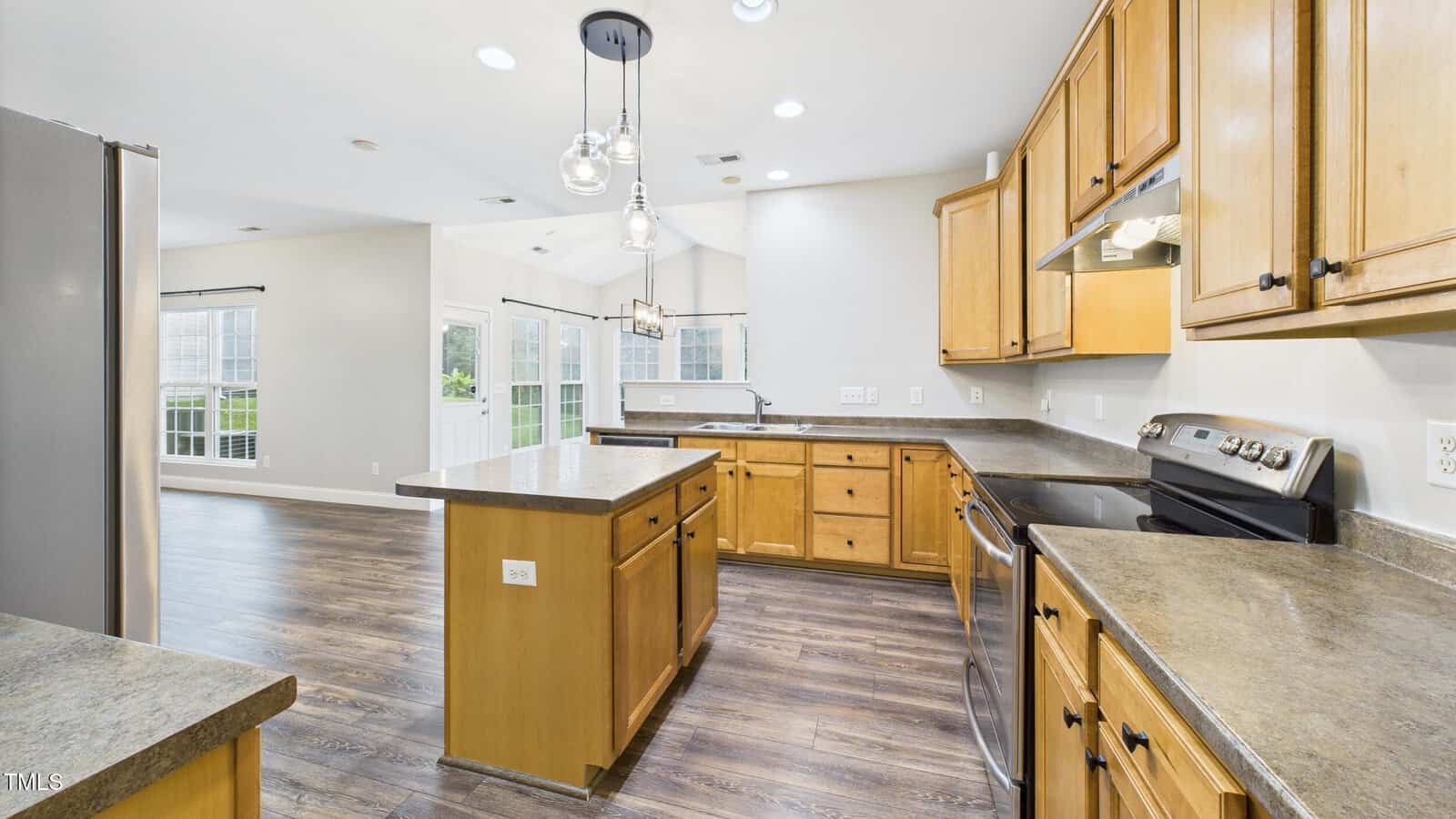 Modern kitchen with wood cabinets, stainless steel appliances, gray countertops, island with storage, pendant lights, and dark wood flooring. Open layout leads to a bright dining area with large windows and a door to the outside.