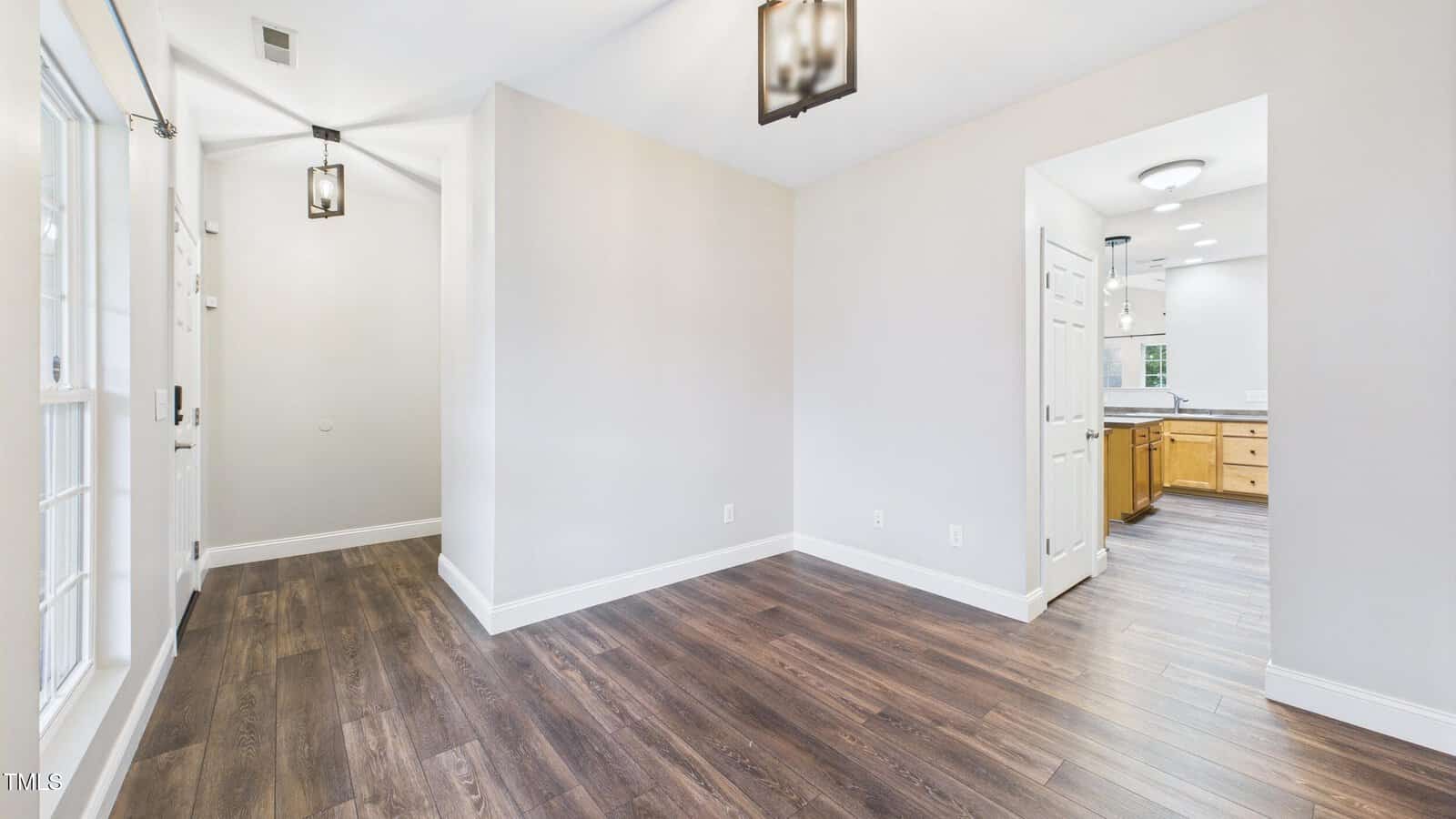 A bright, empty room with white walls and dark wood floors, featuring two ceiling lights, a doorway leading to a kitchen, and a hallway with a door and sidelights.