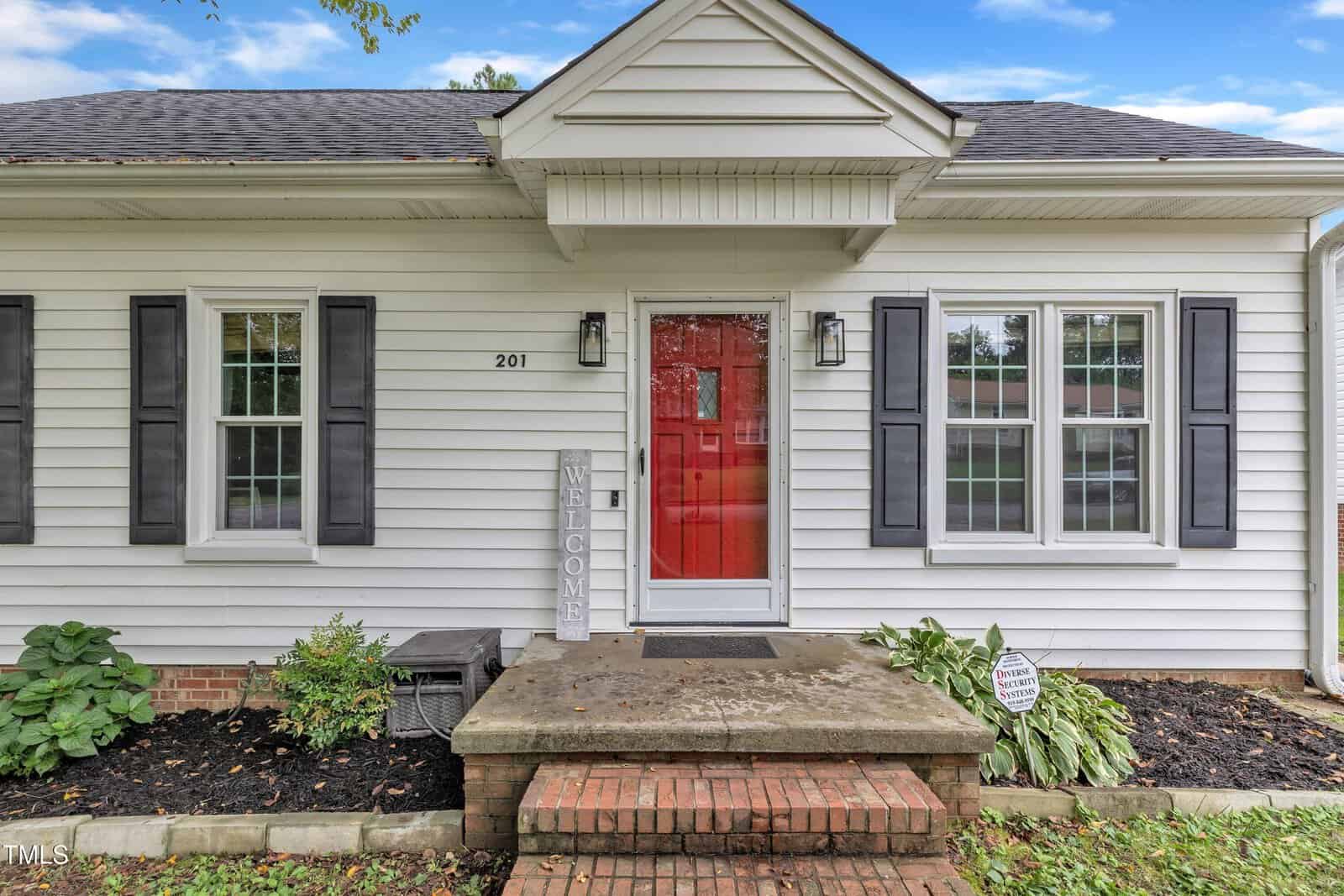 A white house with black shutters, a bright red front door, and a small porch with brick steps. There is a "WELCOME" sign by the door and plants on either side of the entry. House number 201 is visible next to the door.