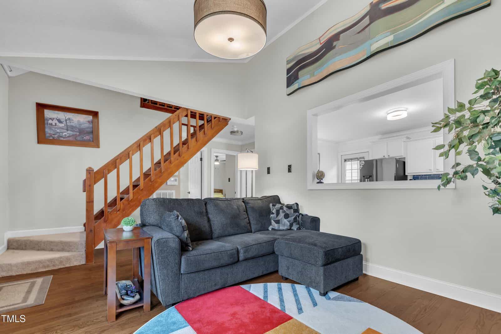 A living room with a dark gray sectional sofa, a colorful round rug, a wooden staircase, a potted plant, wall art, and a pass-through window showing a white kitchen in the background.