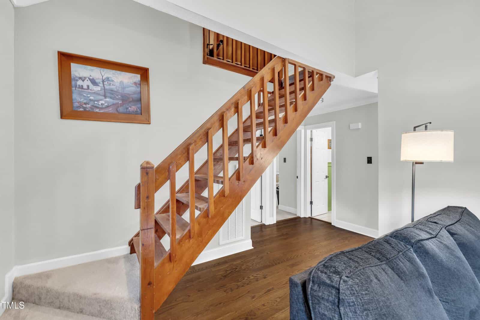 A wooden staircase with open risers leads to an upper level in a room with hardwood floors and light-colored walls. A framed painting hangs on the wall, and a blue couch is partially visible in the foreground.