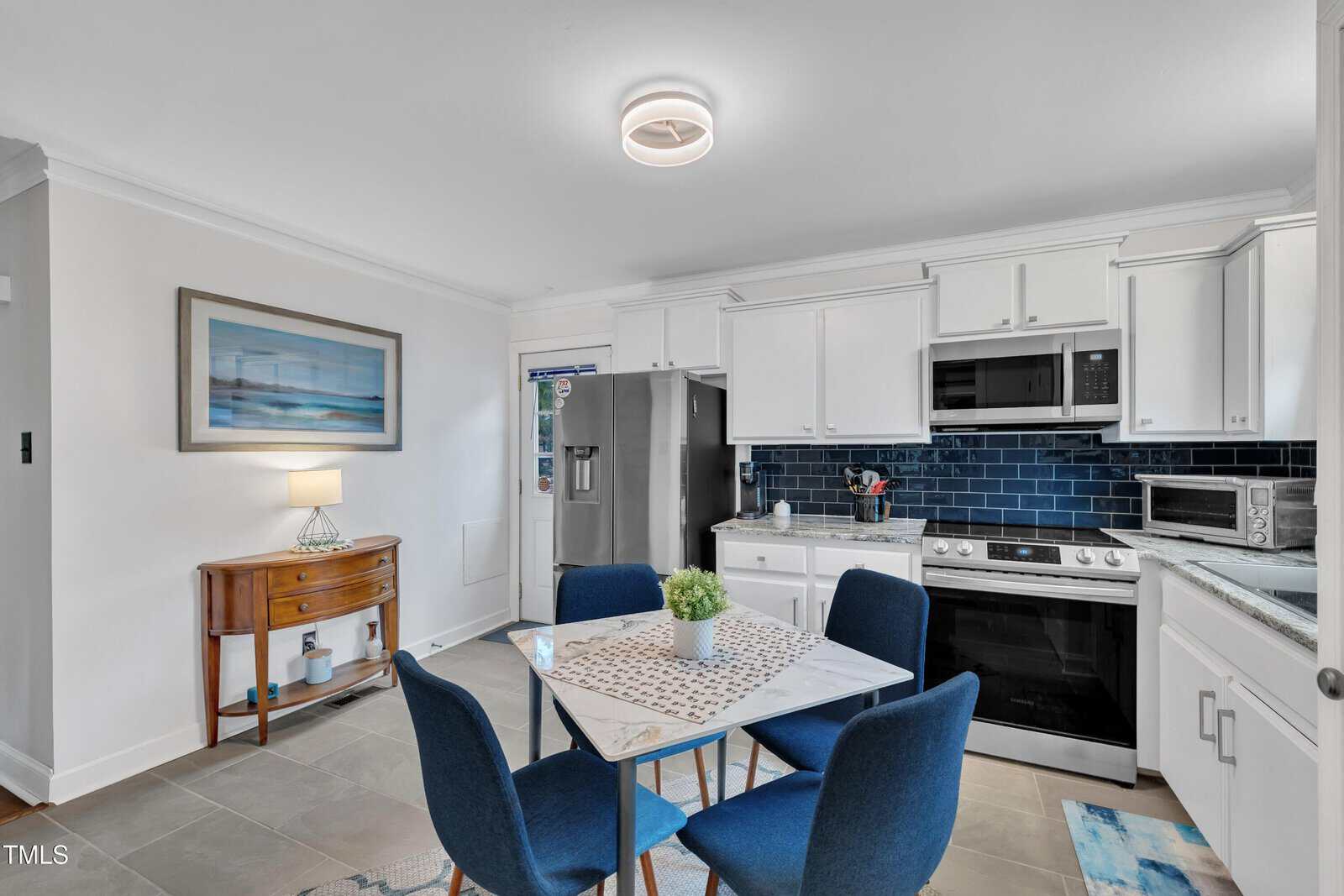 Modern kitchen with white cabinets, stainless steel appliances, blue tile backsplash, a small dining table with four blue chairs, and a side table with a lamp and framed artwork on the wall.