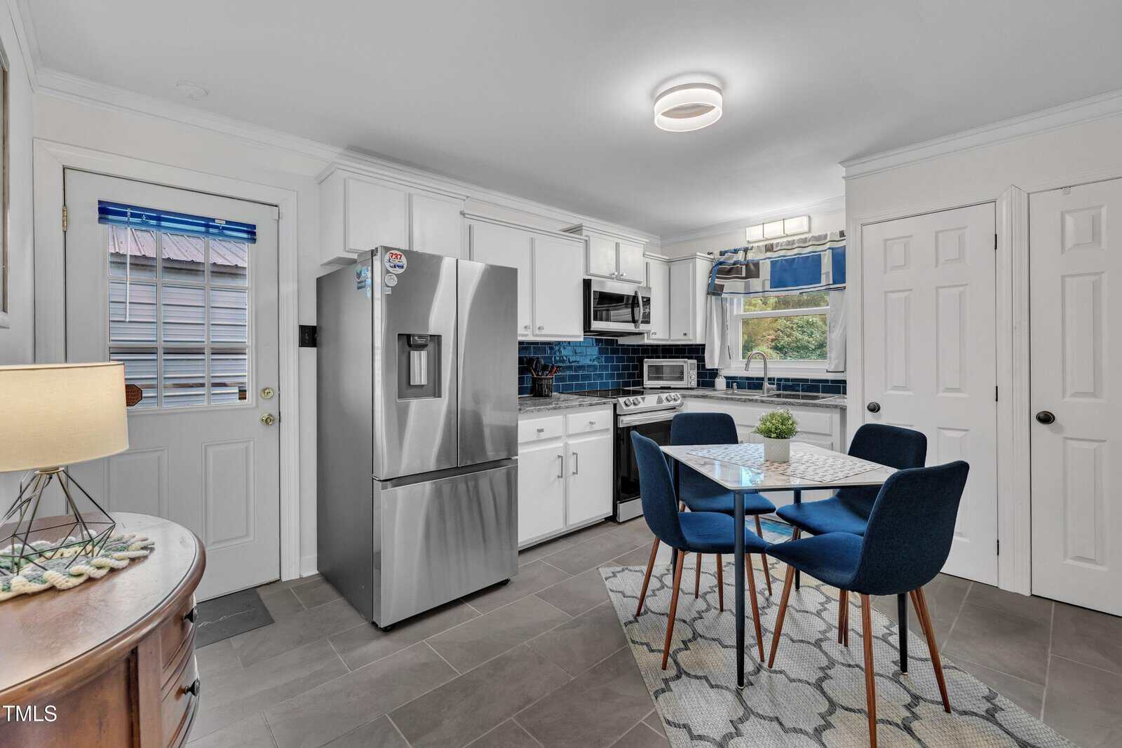 Modern kitchen with stainless steel appliances, white cabinets, blue backsplash, small dining table with four blue chairs, window above sink, and gray tile floor. A lamp and sideboard are partially visible on the left.