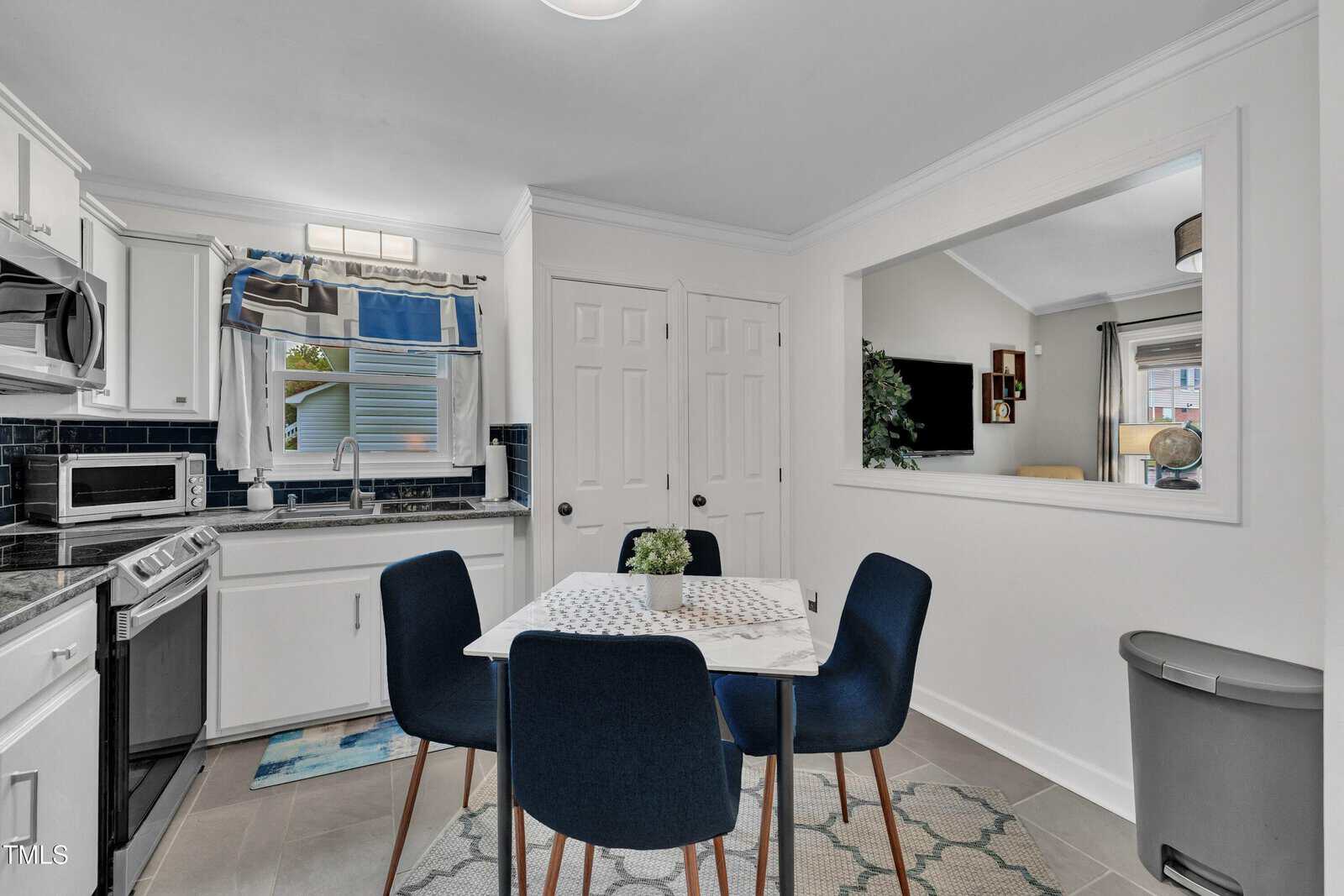 A modern kitchen with white cabinets, stainless steel appliances, a small dining table with four dark blue chairs, and a window above the sink. A large interior window opens to the living room.
