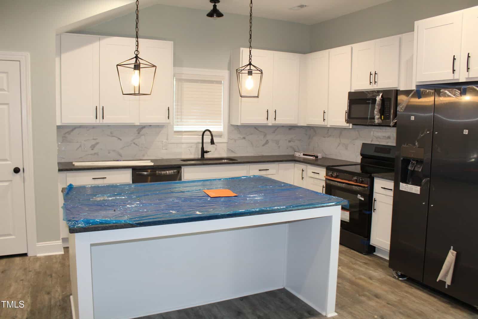 A modern kitchen with white cabinets, stainless steel appliances, marble backsplash, and two pendant lights over a center island. The island is covered in blue protective plastic. A paper sits on the island.