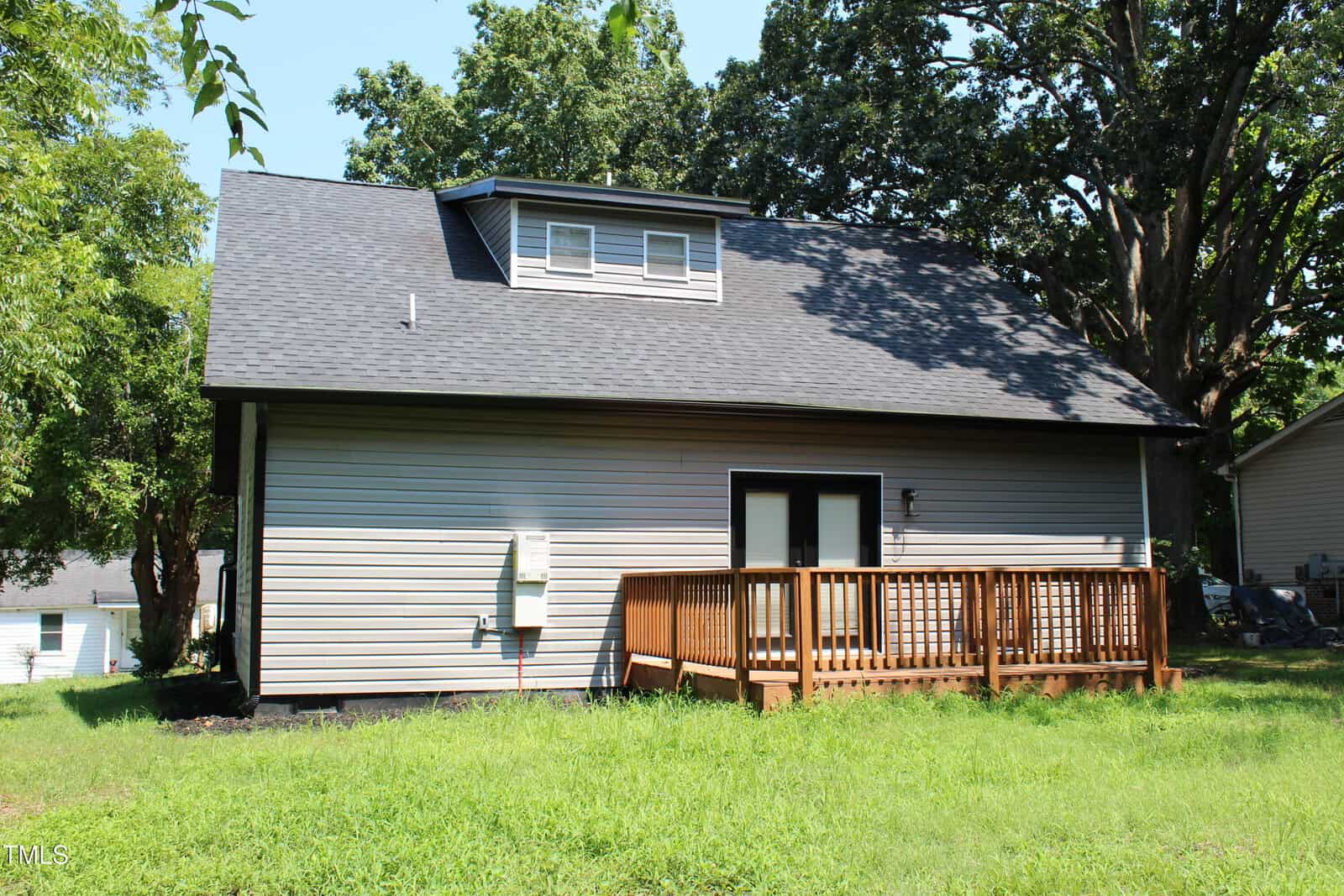 A one-story house with light-colored siding, a dark shingled roof, and a small upper dormer. There is a wooden deck outside a set of glass doors, with grass and trees surrounding the home.