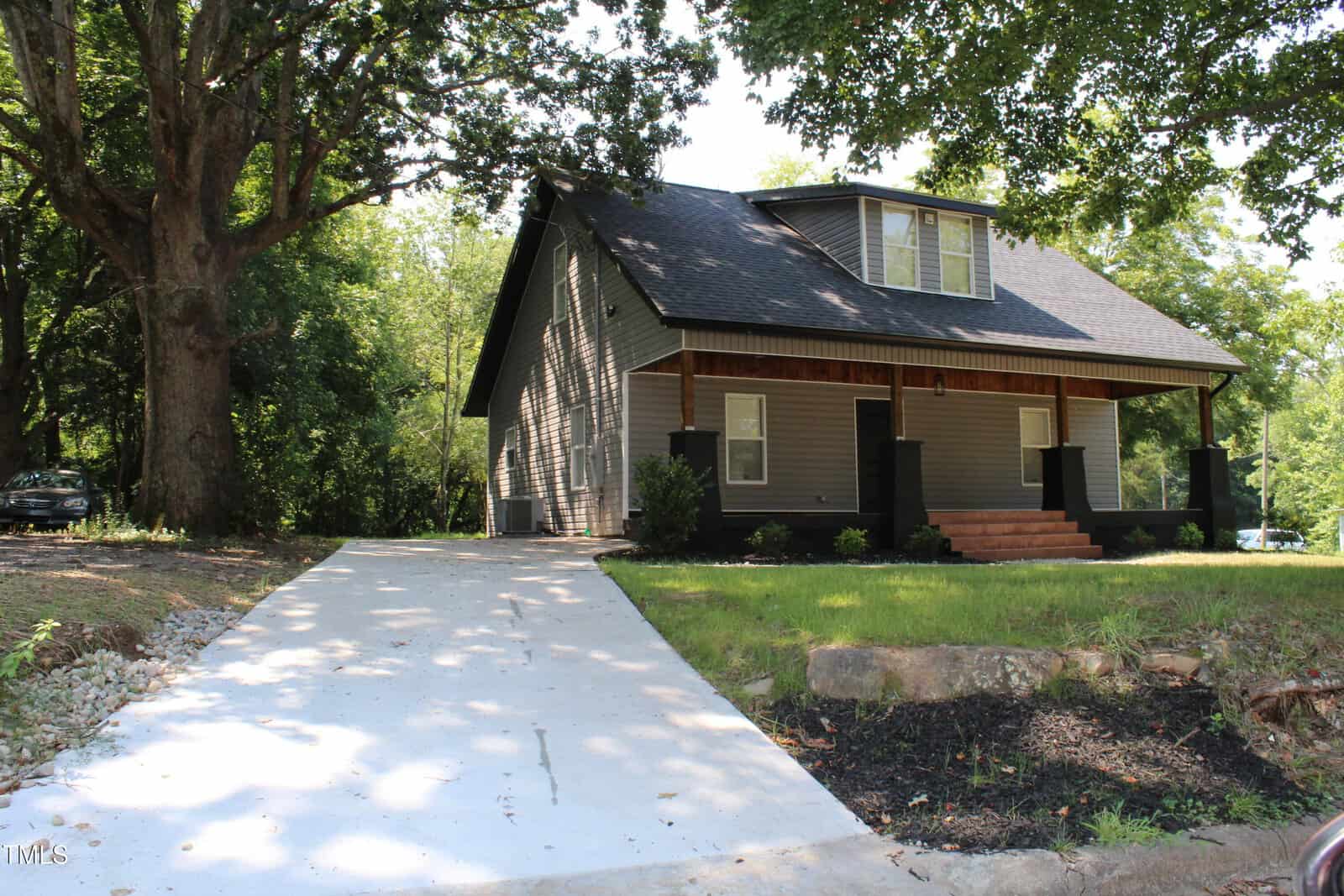 A two-story house with gray siding and a black roof sits next to a large tree. A concrete driveway leads to the front steps. The front porch is covered, and there is landscaping near the entrance.