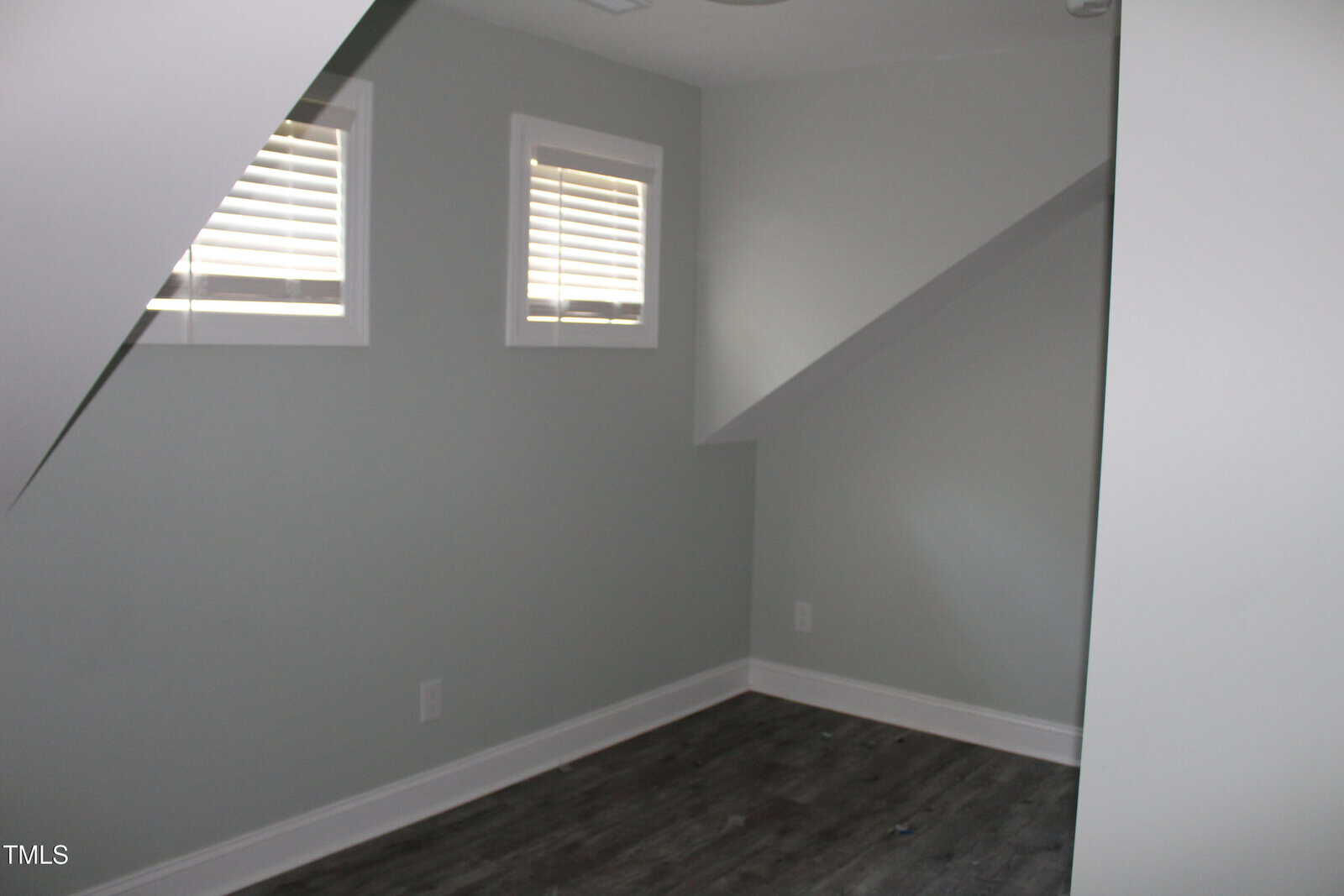 A small, empty room with light gray walls, dark wood flooring, two small windows with blinds, and sloped ceilings creating angular shapes on the left and right sides.