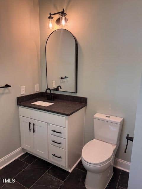 Modern bathroom with a white vanity, black countertop, oval mirror, black fixtures, and a white toilet. Overhead light fixture and dark tile floor are also visible.