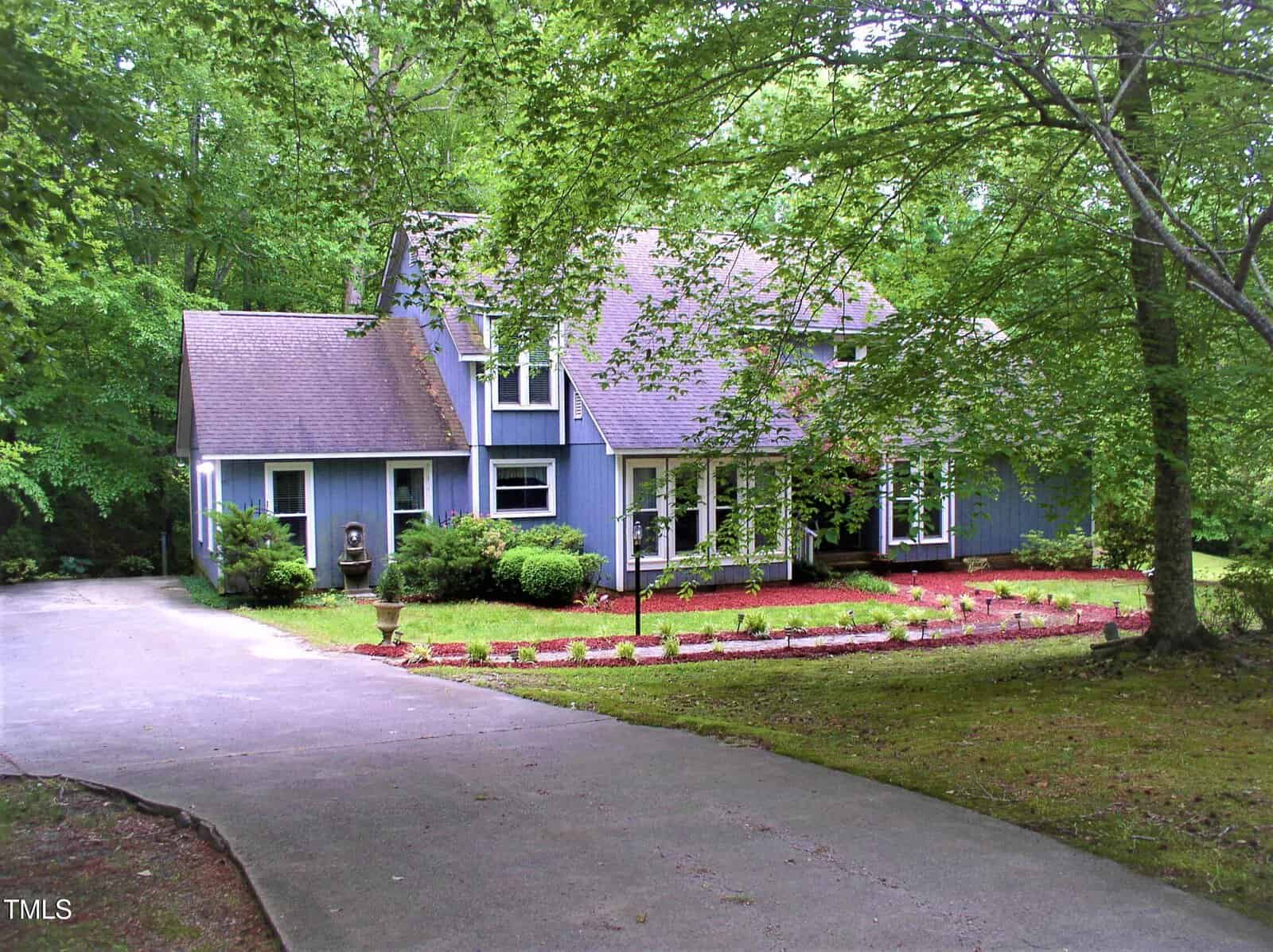 A two-story blue house with white trim is surrounded by trees and greenery. A curved concrete driveway leads to the front of the house, with landscaped beds and a well-maintained lawn in the yard.