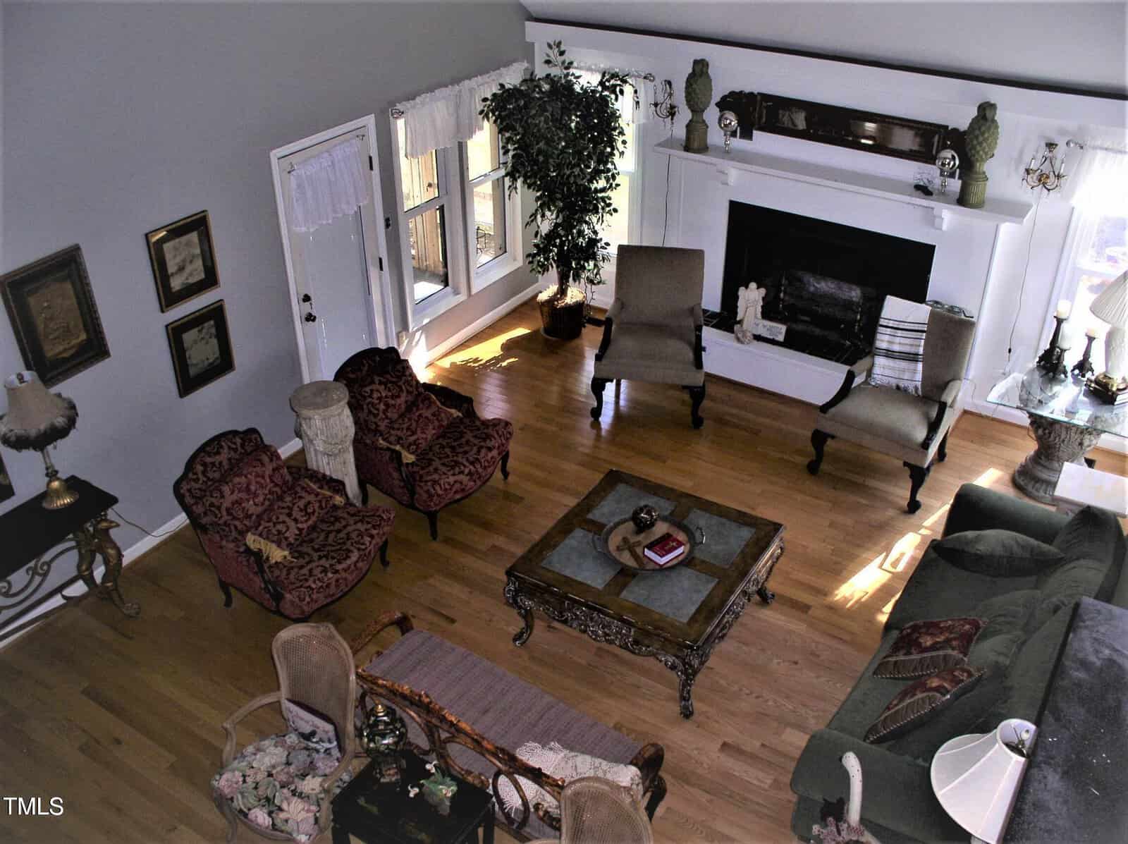 A living room with hardwood floors, a fireplace, several chairs, a sectional sofa, a central coffee table, a side table with a lamp, framed pictures on the wall, and a potted plant near a window.