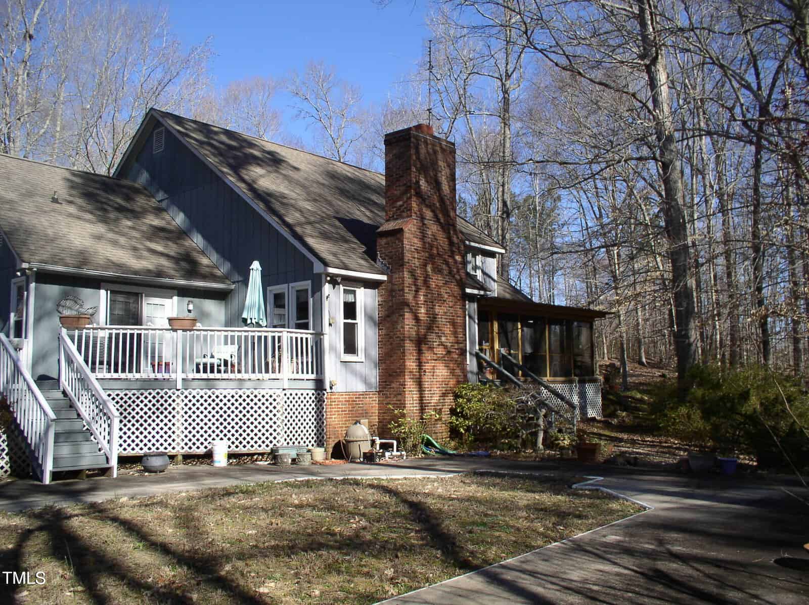 A two-story gray house with a brick chimney features a raised deck with white railings and a screened porch. Leafless trees surround the house, and there are potted plants and a paved walkway in the yard.