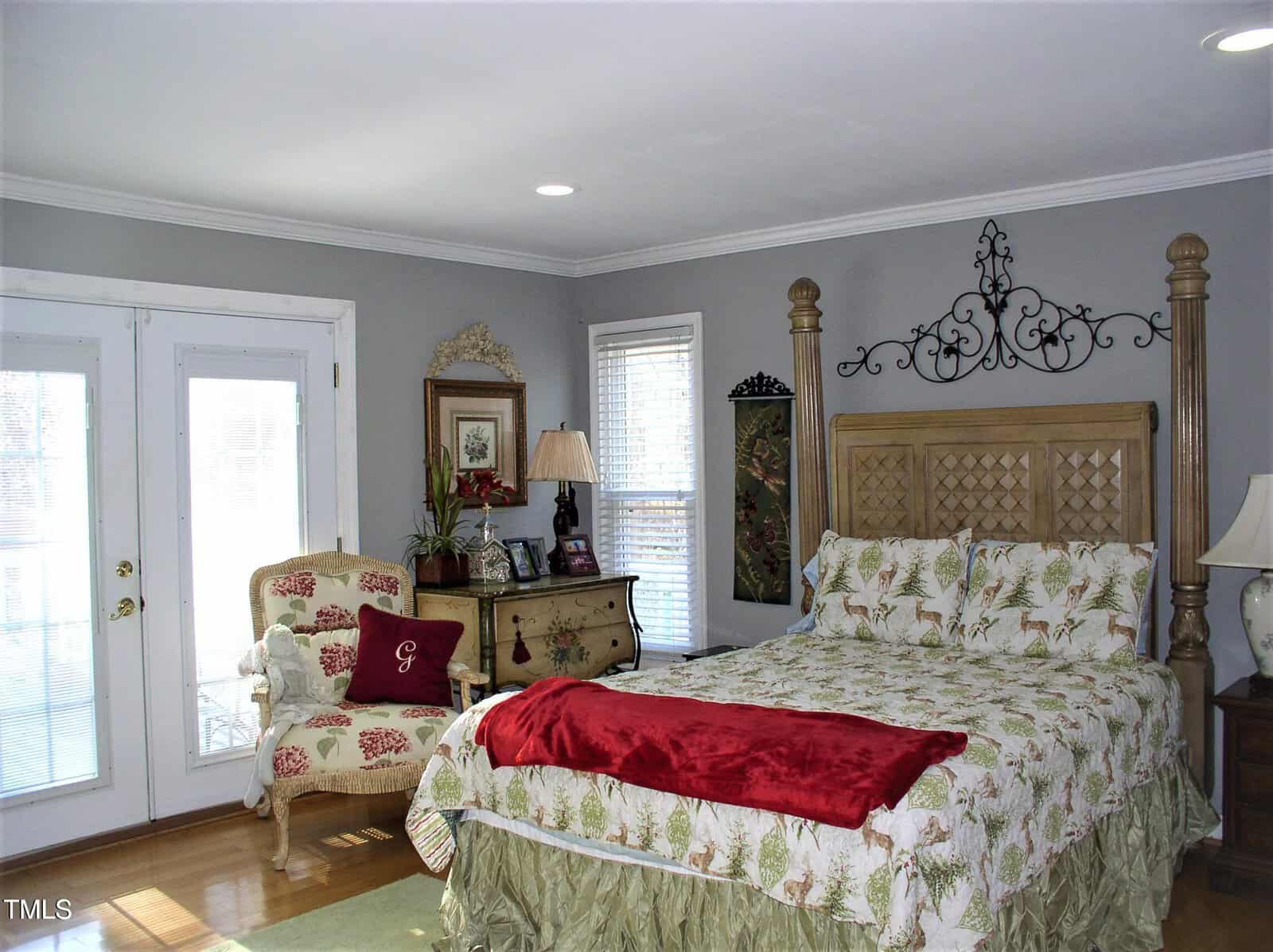 A bedroom with a large wooden bed, floral bedding, and a red throw blanket. There is an upholstered chair and a dresser with a lamp. French doors and two windows let in natural light. The walls are painted light gray.