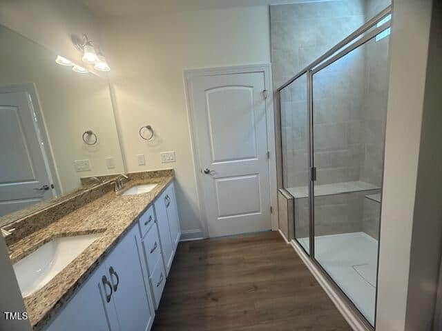 Modern bathroom with double sinks on a granite countertop, white cabinets, large mirror, wood-look flooring, and a glass-enclosed shower with beige tiles and a built-in bench. White door on the back wall.