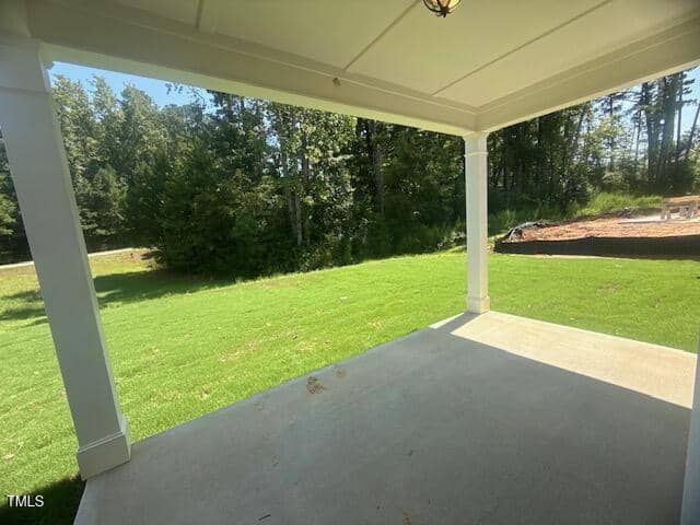 Covered patio with a concrete floor overlooks a grassy backyard bordered by trees. Sunlight casts shadows on the lawn and part of the patio. A pile of dirt or mulch is visible off to the right.