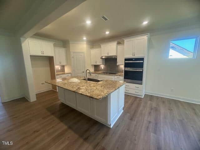 Modern kitchen with white cabinets, granite island countertop with sink, built-in ovens, tile backsplash, wood flooring, and recessed lighting. There is a small window and an empty space for a refrigerator.