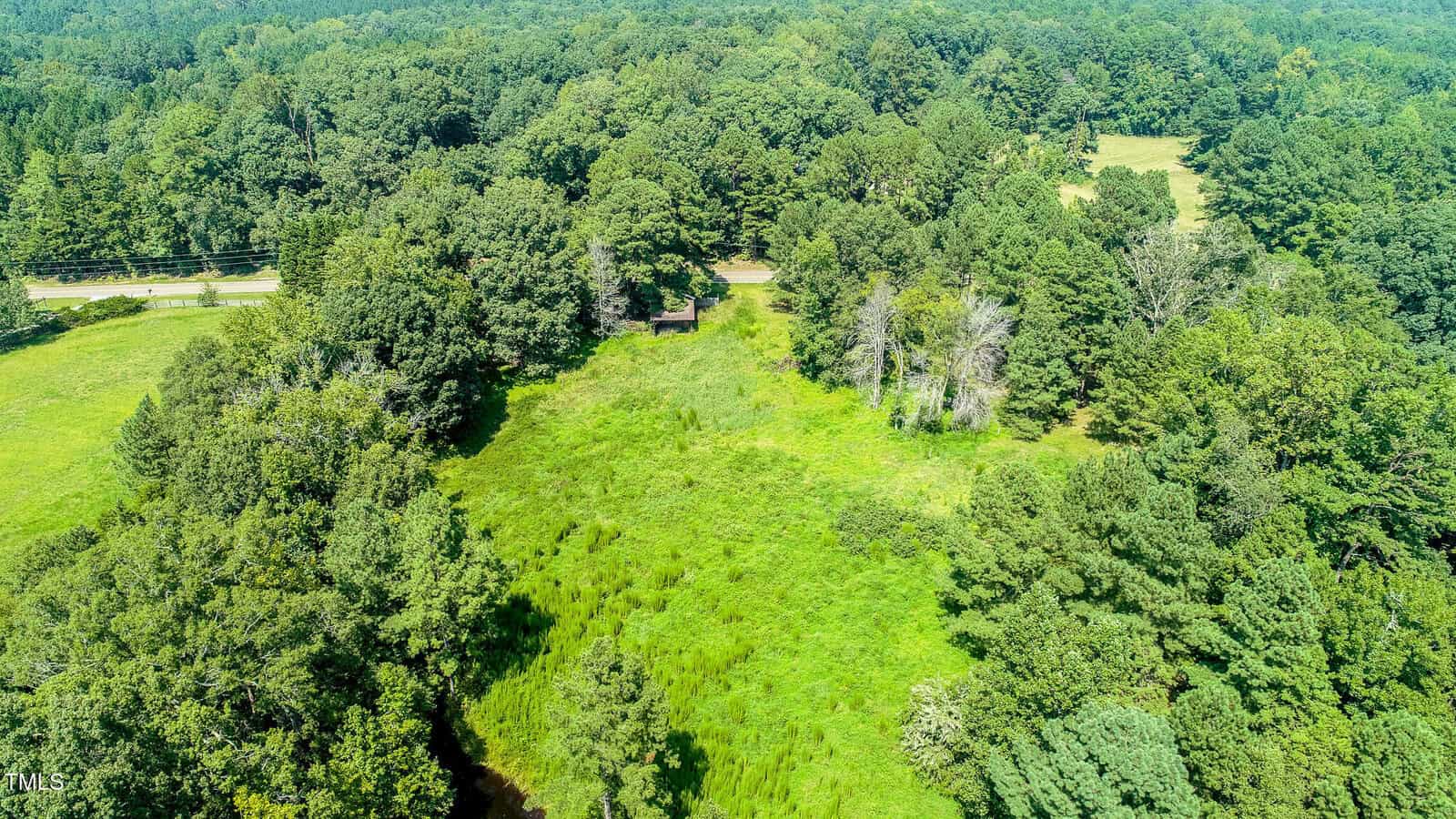 Aerial view of a green, grassy clearing surrounded by dense trees, with a small building near the center and a road visible at the top edge of the image.