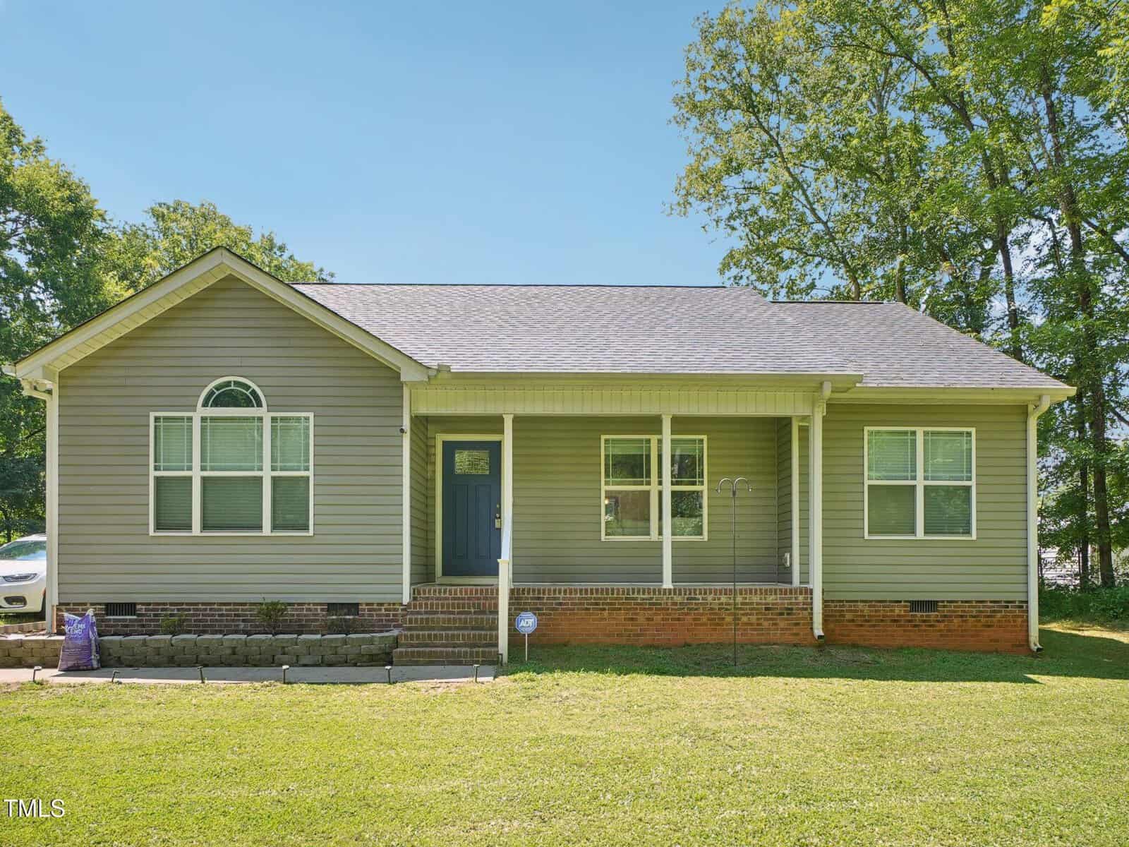 Charming single-story house at 2164 Smith Road, Oxford, featuring light gray siding, a dark shingled roof, white trim, a covered front porch, brick foundation, and a grassy front yard with trees in the background.