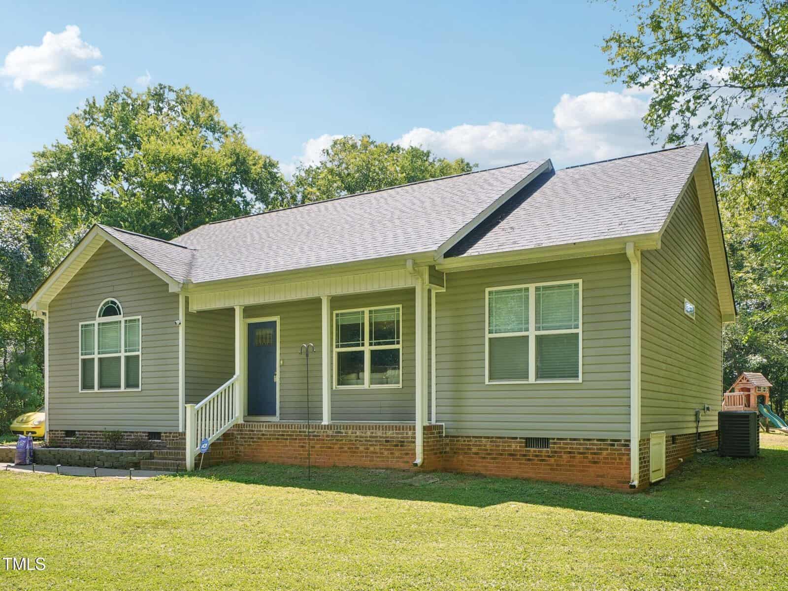 A single-story house with gray siding, a covered front porch, white trim, and a brick foundation. The yard is grassy with a few trees in the background and a playset visible to the right.