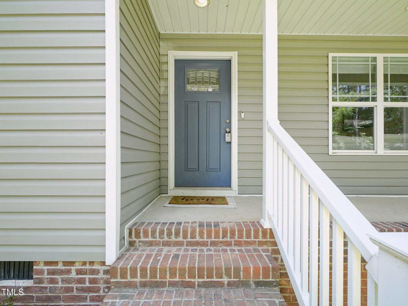 A front porch with brick steps, a blue door with a silver handle and lock, beige siding, white trim, a white railing, and a doormat in front of the door. A window with white frames is to the right of the door.
