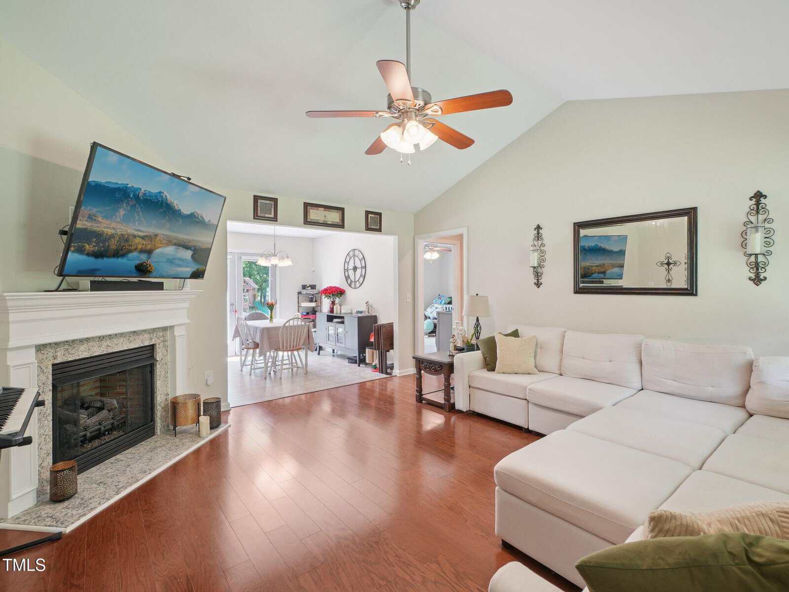 A living room with hardwood floors, a white sectional sofa, a wall-mounted TV above a fireplace, ceiling fan with lights, and wall art. The room connects to a dining area through an open doorway.