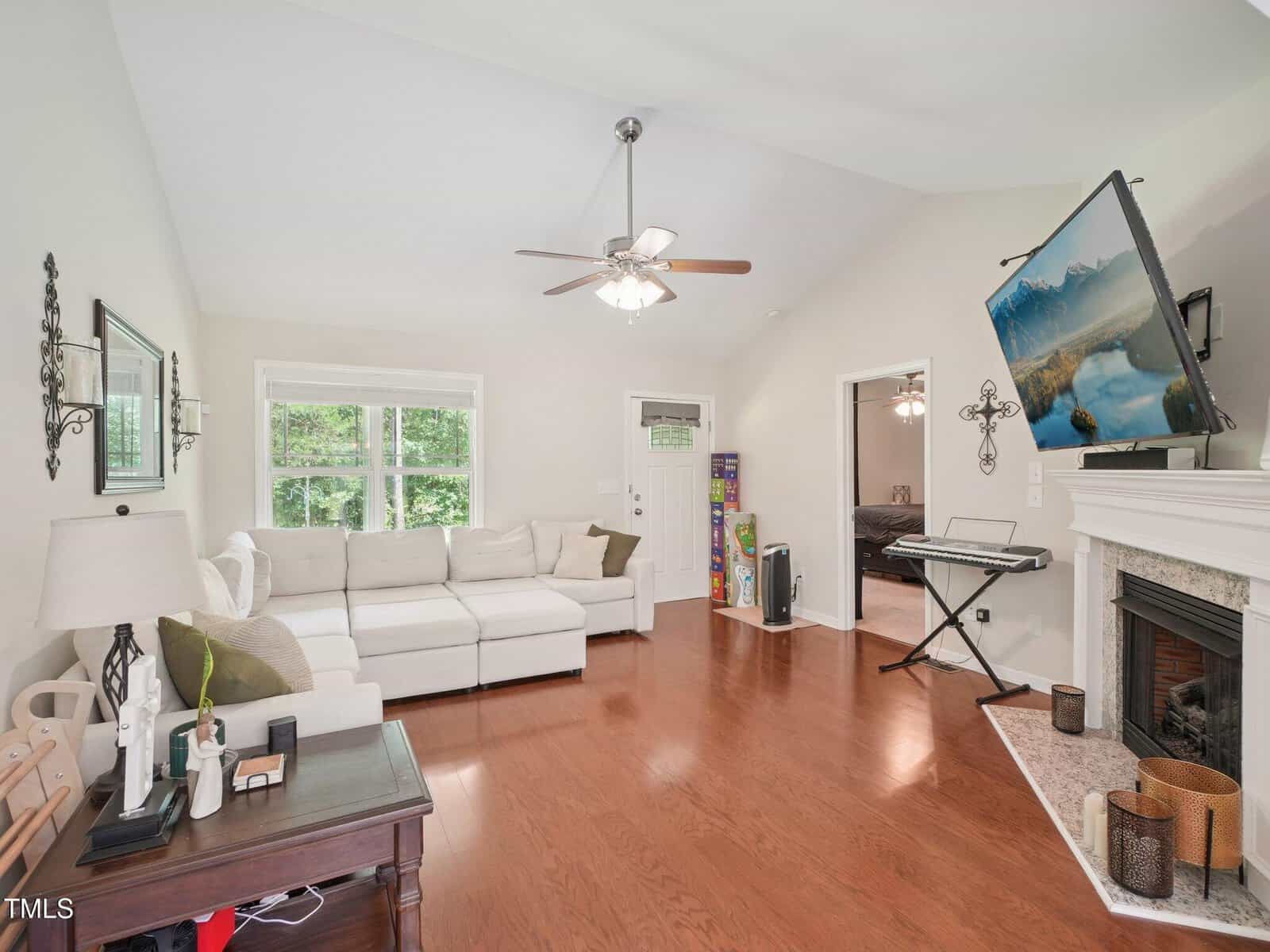 A bright living room with a white sectional sofa, a dark side table, a fireplace with candles, a TV mounted above the mantel, an electronic keyboard, and large windows letting in natural light.
