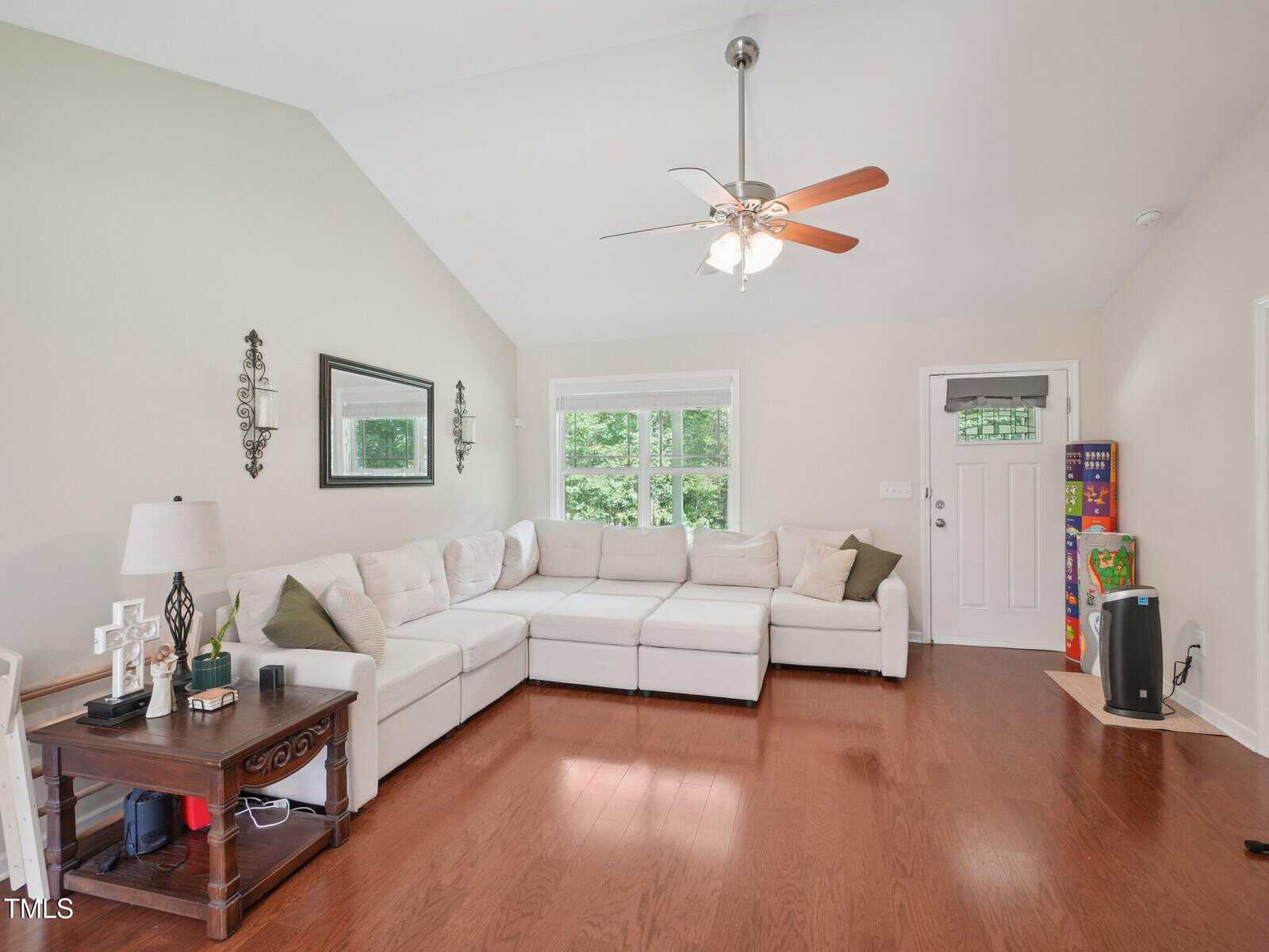 A spacious living room with a white sectional sofa, hardwood floors, a ceiling fan with lights, side table, wall mirror, and two wall sconces. Large windows and a door provide natural light; a stack of games is in the corner.