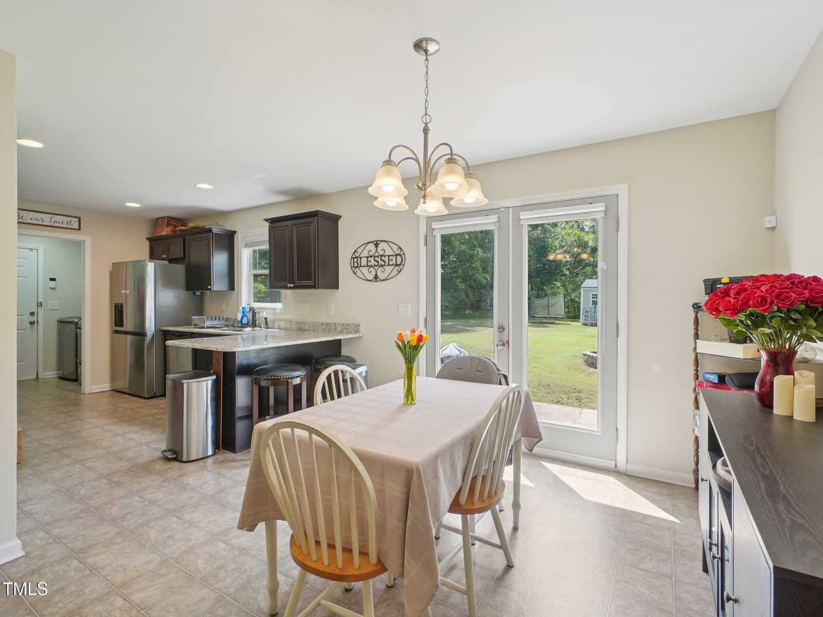A bright kitchen and dining area with a table for four, flowers in a vase, dark cabinets, stainless steel appliances, and glass doors leading to a backyard with green grass and trees.