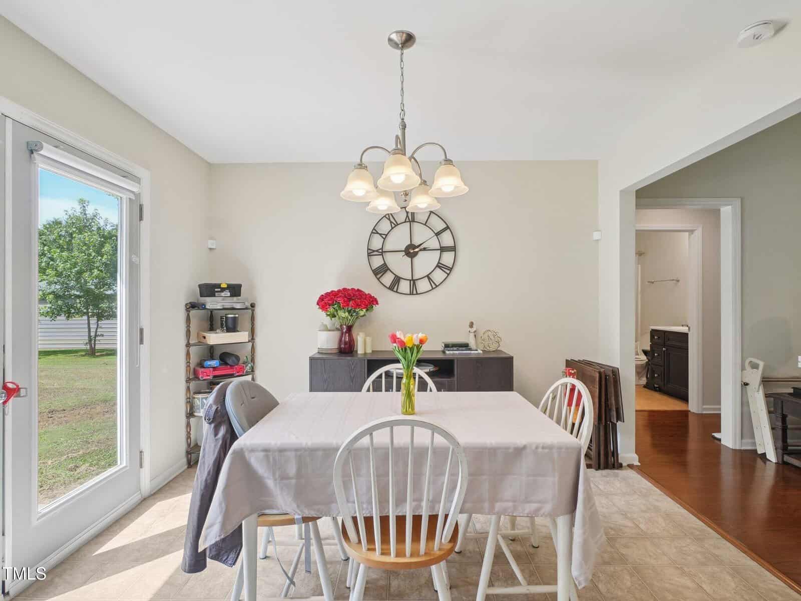 A dining area with a rectangular table covered by a gray tablecloth, four chairs, a vase of tulips, a sideboard with flowers, a large wall clock, and a sliding door showing a view of the backyard.