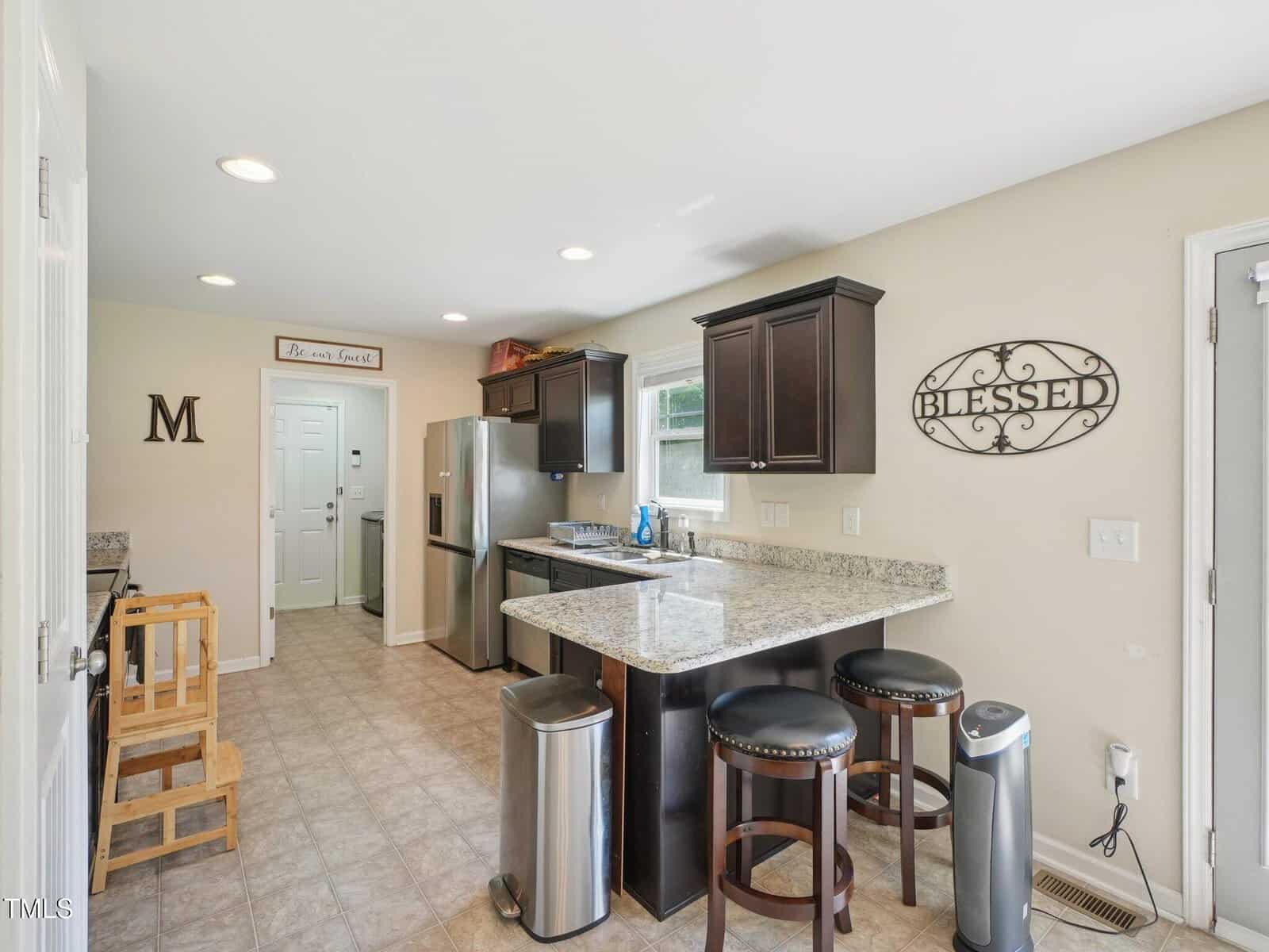 A kitchen with beige walls, dark cabinets, stainless steel appliances, granite countertops, and a breakfast bar with two stools. Wall decor includes a metal "Blessed" sign and a letter "M.