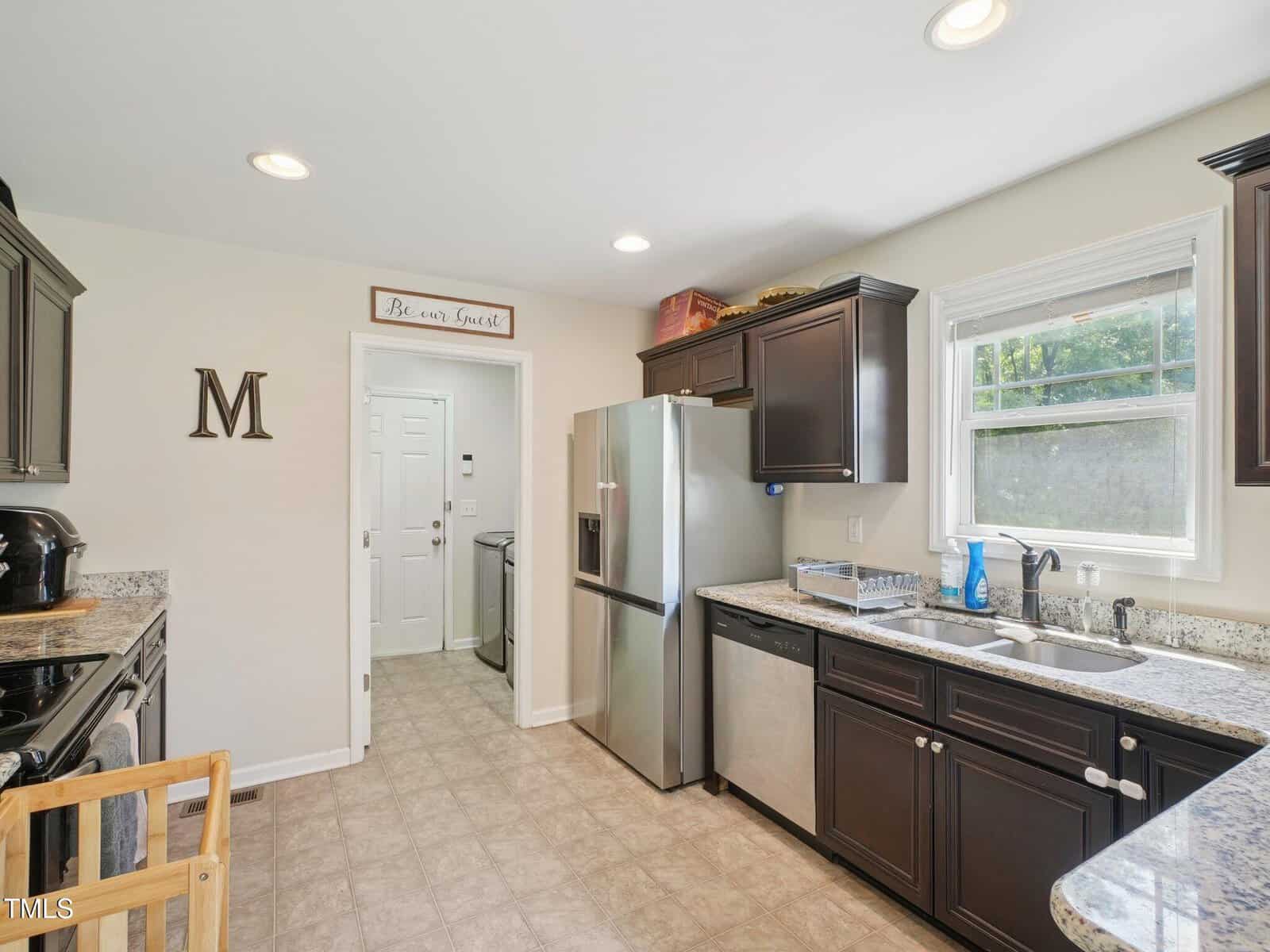 A kitchen with dark cabinets, stainless steel appliances, granite countertops, a double sink under a window, and a view into a laundry room. A wooden letter "M" and sign hang on the wall.