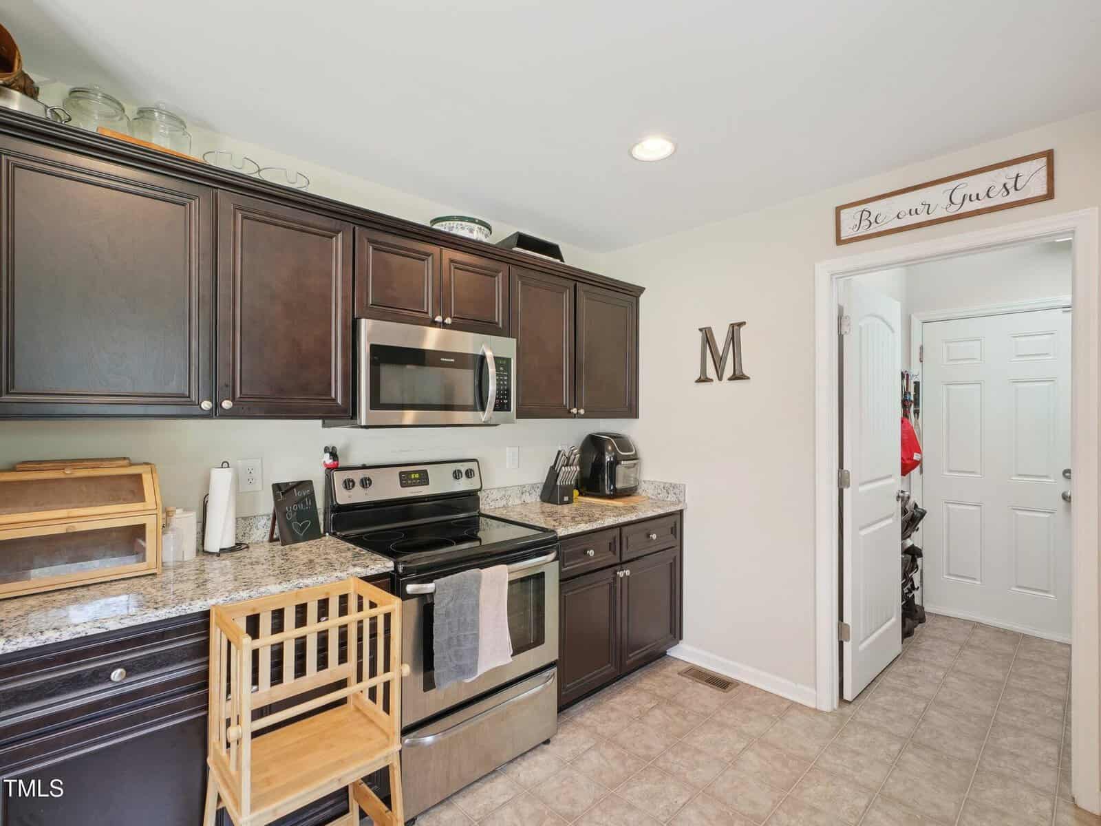 A kitchen with dark cabinets, stainless steel appliances, a wooden high chair, and granite countertops. A “Be our Guest” sign is above the doorway leading to a pantry or laundry area.