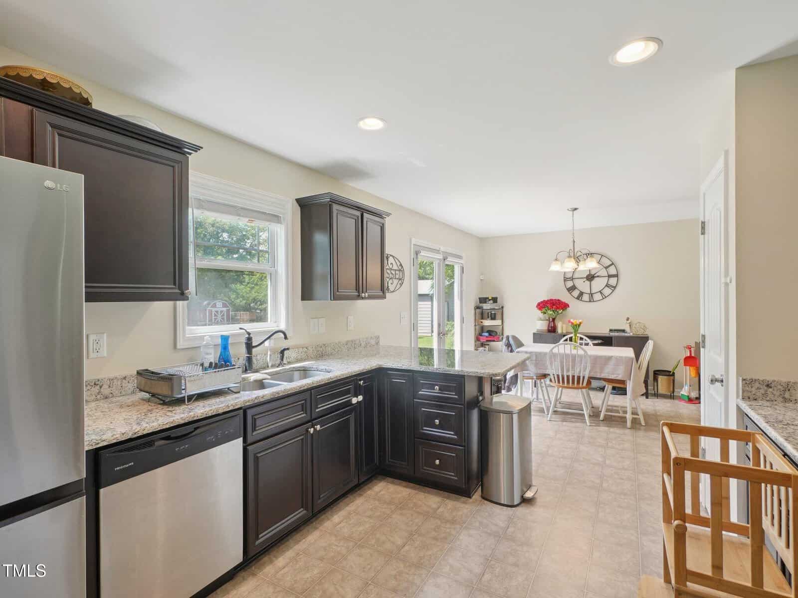 Bright kitchen with dark cabinets, stainless steel appliances, and granite countertops. Dining area with table and chairs is visible in the background, with large windows letting in natural light.