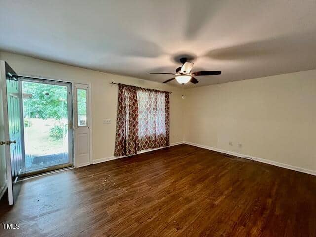 Empty living room with wood flooring, a ceiling fan with lights, a large window with patterned curtains, and an open door leading outside; walls are painted a light color.