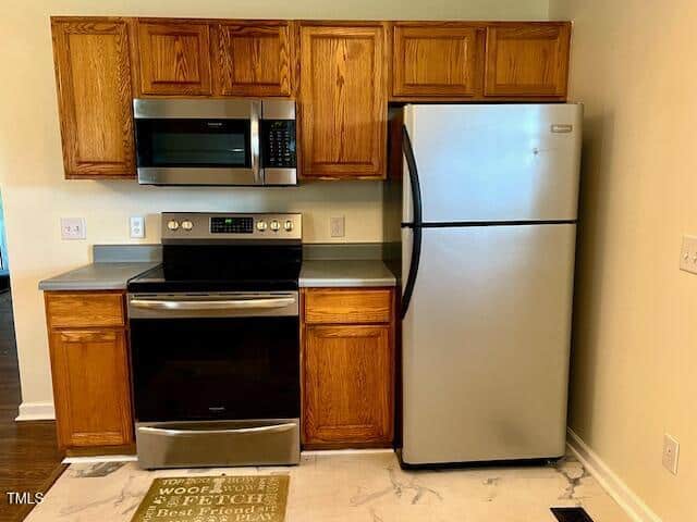 A kitchen with wooden cabinets, stainless steel microwave, electric stove, and refrigerator. Light countertops and a rug with text are on the floor in front of the appliances.
