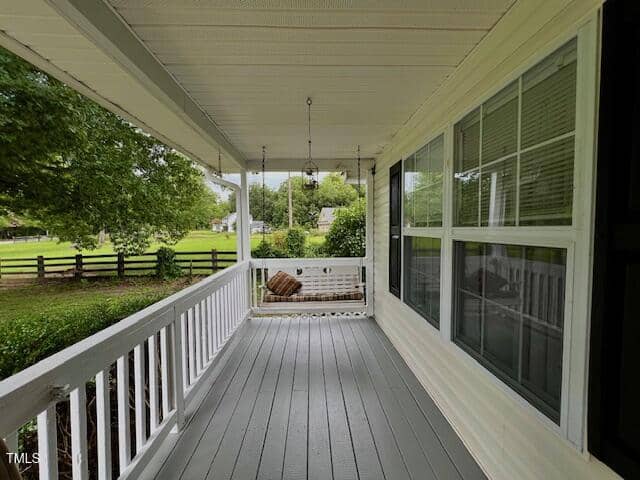 A covered front porch with gray flooring, white railings, and large windows on the right. A ceiling fan and hanging light fixture are overhead. Trees, a wooden fence, and grass are visible in the background.