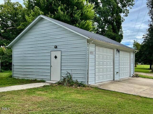 A white metal garage with a single side door and two garage doors stands on a concrete driveway surrounded by green grass and trees. The building has a gray metal roof and a small light above the side door.