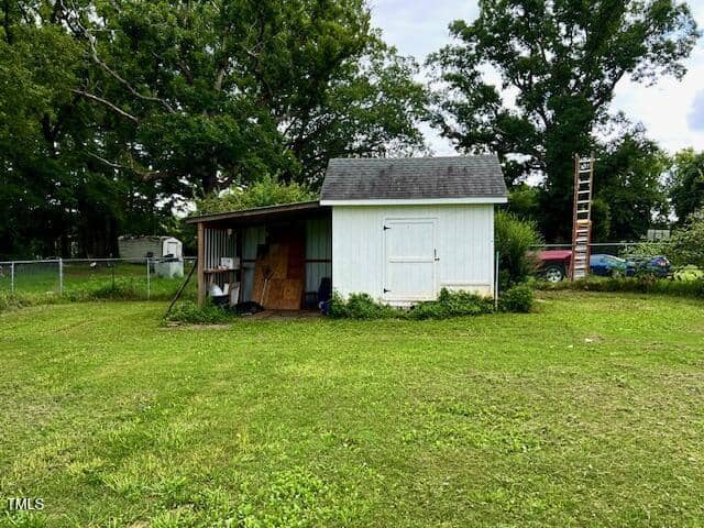 A small white shed with a gray roof stands on a grassy lawn. The shed has double doors, an open lean-to area with various items stored inside, and is surrounded by trees and a chain-link fence. A ladder leans on the right side.