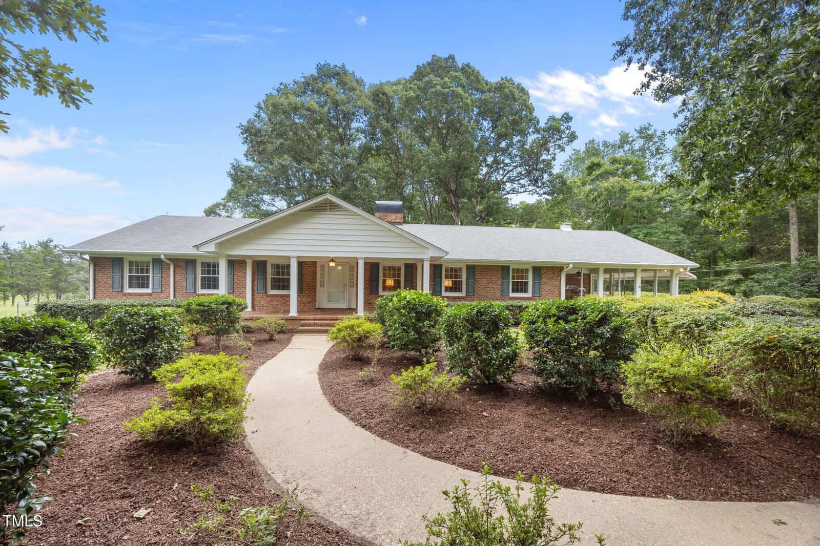 This charming single-story brick house at 5626 Tabbs Creek Road, Oxford, features white trim and a covered front porch. A curved concrete walkway winds through landscaped bushes to the entrance, framed by large trees and blue sky in the background.