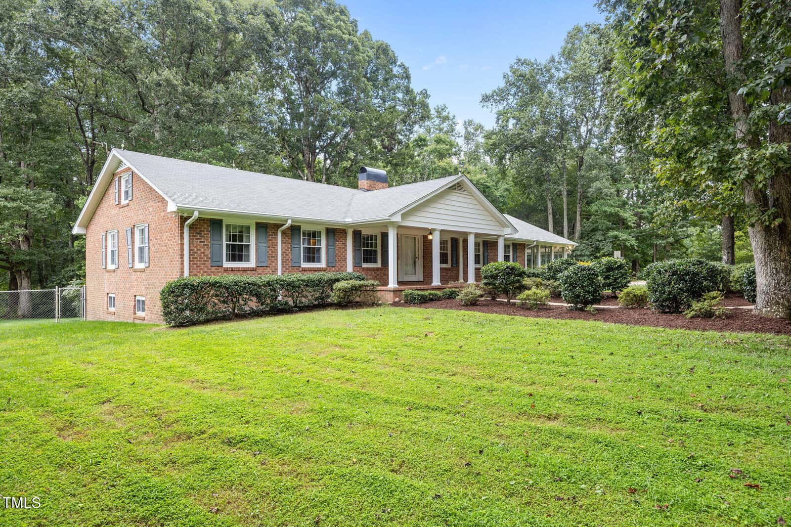 Single-story brick house with a gray roof, white trim, and multiple front windows, surrounded by a well-maintained lawn, shrubs, and mature trees on a clear day.