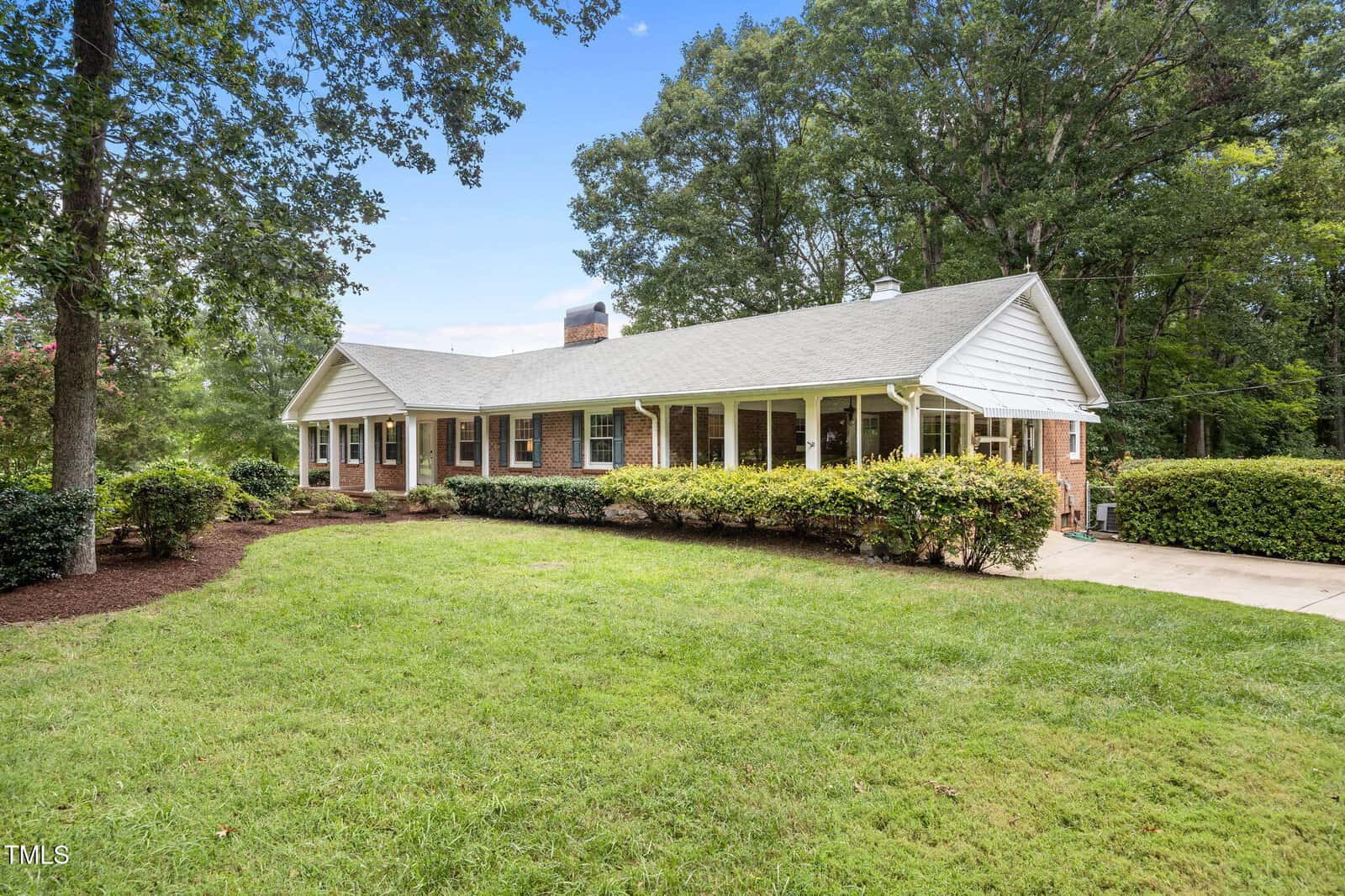 A single-story brick house with white columns and trim, surrounded by well-kept shrubs and a green lawn. Large trees provide shade, and a paved driveway is visible on the right side.