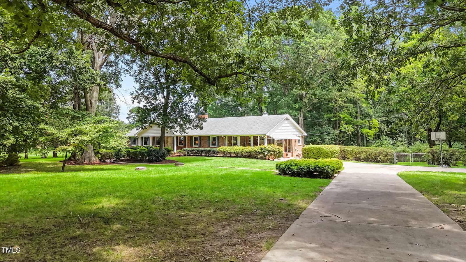 Single-story brick house with white trim and a gabled roof, surrounded by large trees and green lawn, with a driveway curving to the right and a wooded area in the background.