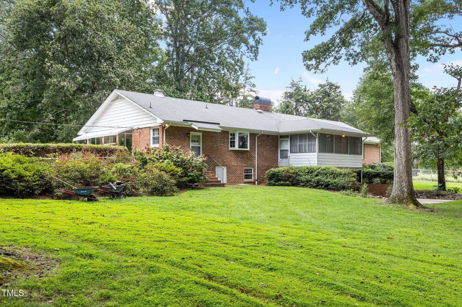 A single-story brick house with a gray roof is surrounded by green grass, shrubs, and mature trees. The house features a screened porch, steps leading to the yard, and a wheelbarrow near the bushes.