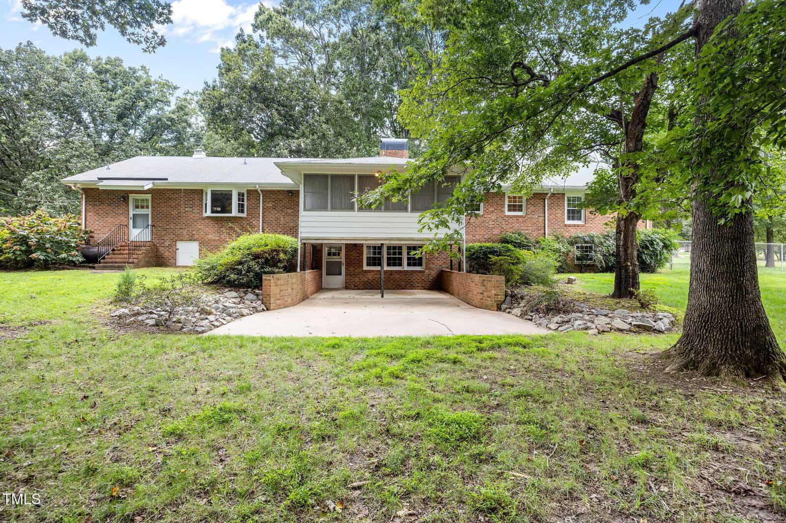 A brick single-story house with a raised screened-in porch, concrete patio, and landscaped garden beds. Large trees and a grassy yard surround the home.