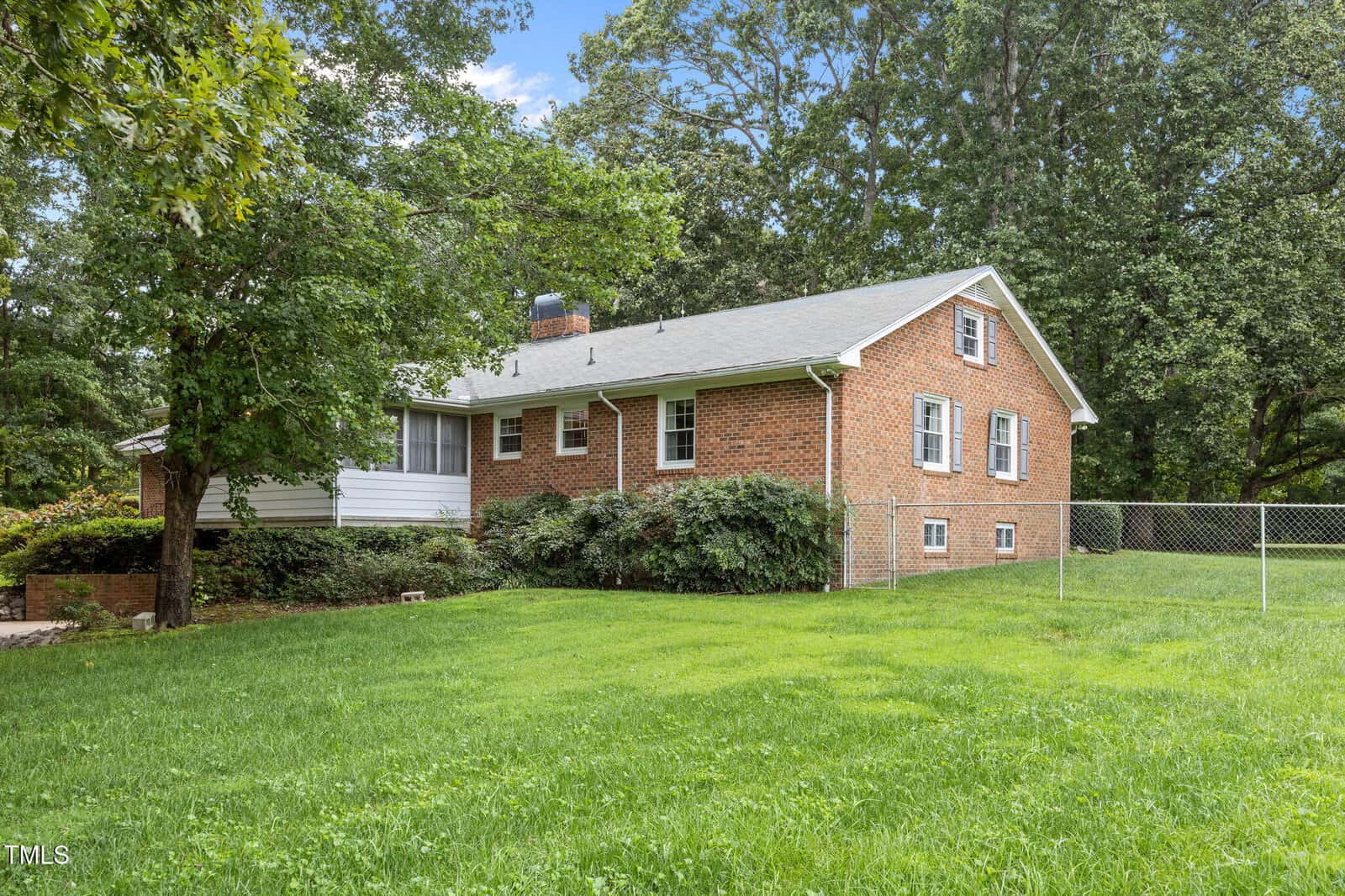 A one-story red brick house with white trim and a gray shingle roof is surrounded by green grass, bushes, and large trees. A chain-link fence runs along the side yard. The sky is blue with scattered clouds.