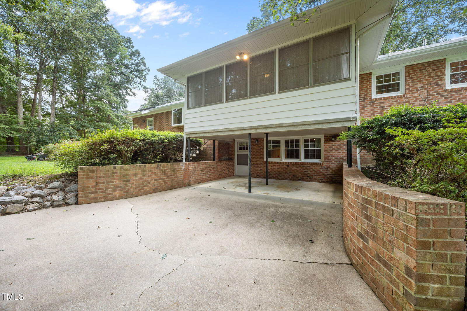 A brick house with a large concrete driveway, a covered area beneath an enclosed upstairs porch, and surrounding bushes and trees under a partly cloudy sky.