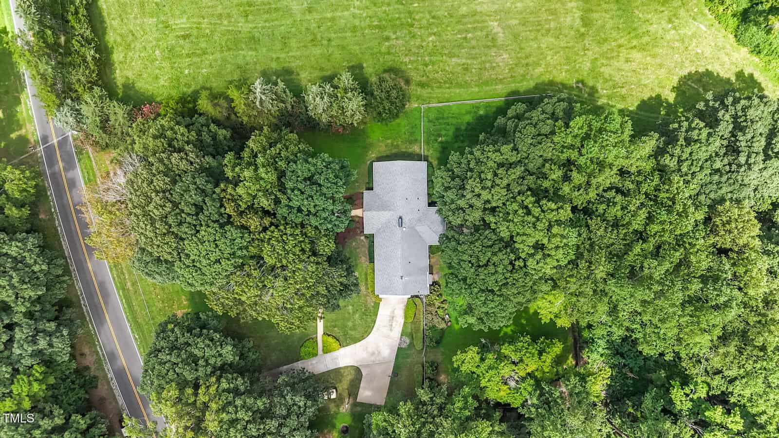 Aerial view of a house with a gray roof surrounded by trees and a large grassy yard. A curved driveway leads from the road to the house, and the property is bordered by more trees and open fields.