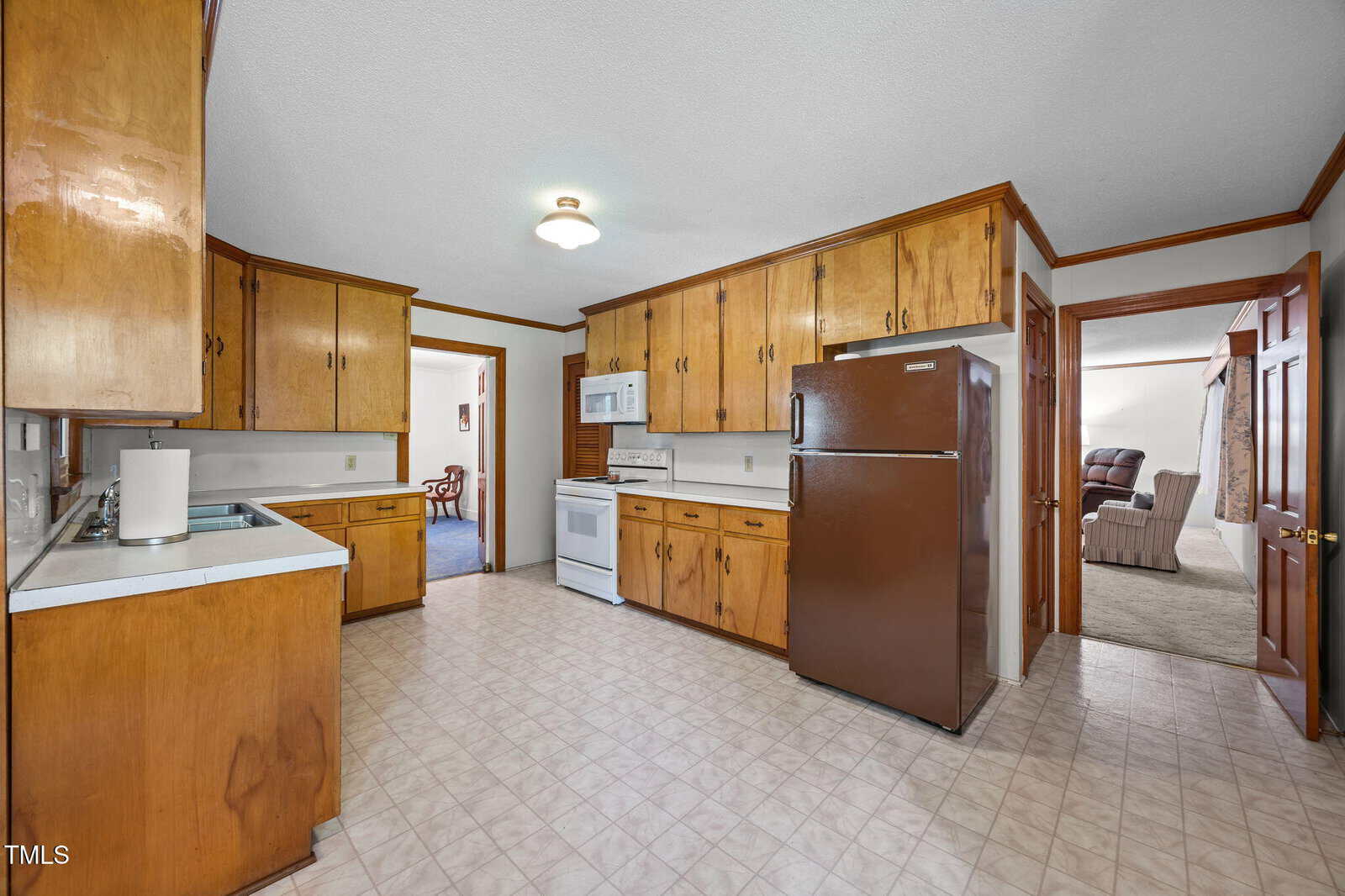 A kitchen with brown wooden cabinets, a brown refrigerator, a white stove, a white microwave, and a beige tile floor. The kitchen connects to a carpeted room on the right and another room in the background.