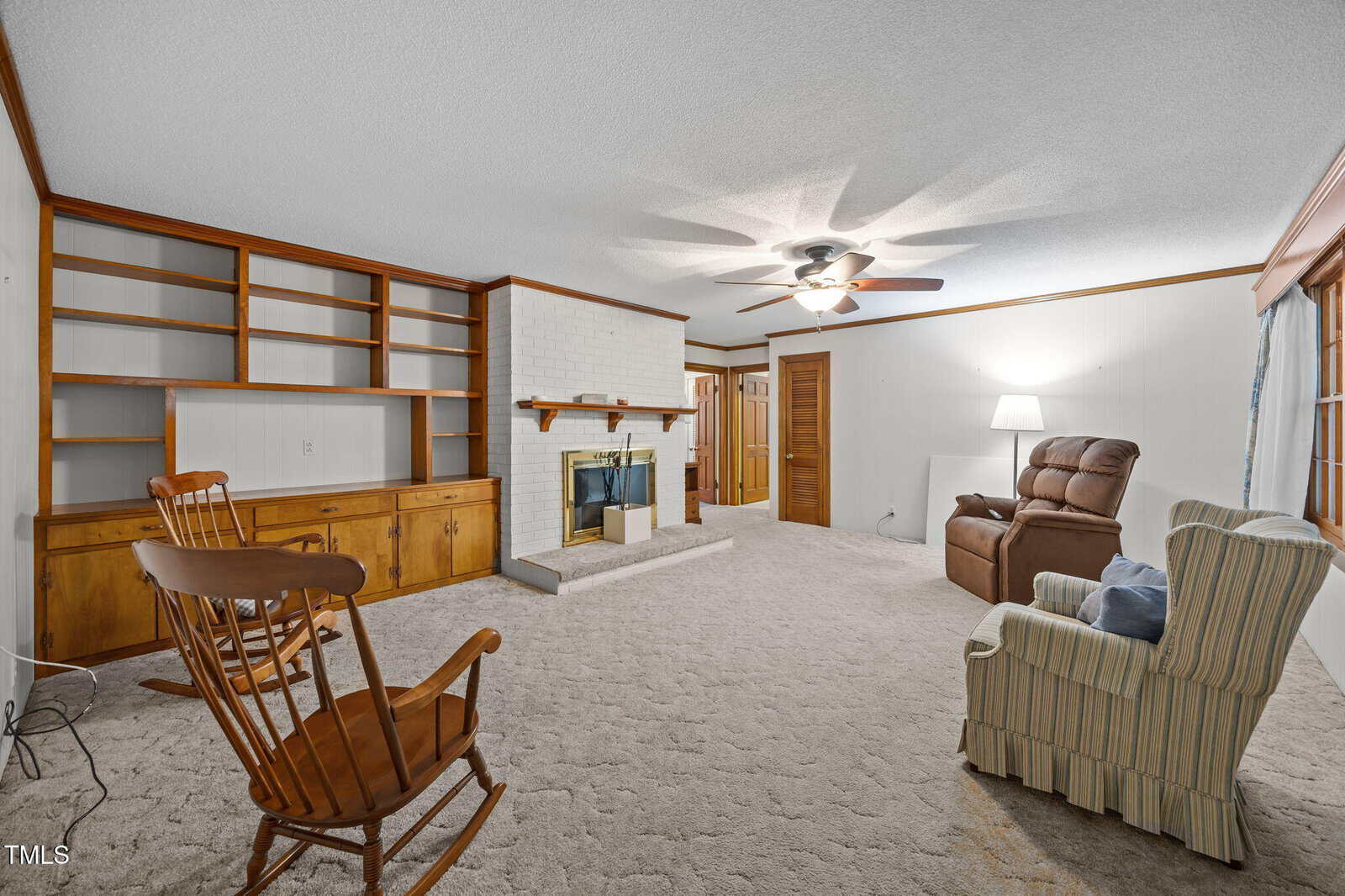 A carpeted living room with a rocking chair, armchair, recliner, built-in shelves, a brick fireplace, ceiling fan, floor lamp, and wood trim along the walls and door.